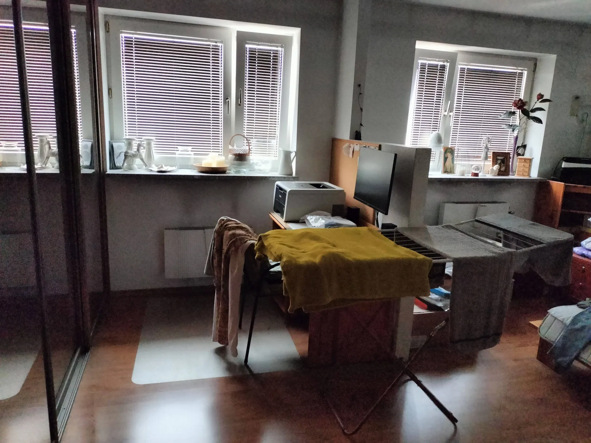 Wnętrze pokoju z dwoma oknami z białymi żaluzjami. W pomieszczeniu znajduje się biurko z monitorem komputerowym i drukarką. Na pierwszym planie stoi rozkładana deska do prasowania przykryta żółtym materiałem, obok której leży brązowy koc. W tle widoczne są przedmioty dekoracyjne na parapetach oraz fragmenty mebli.