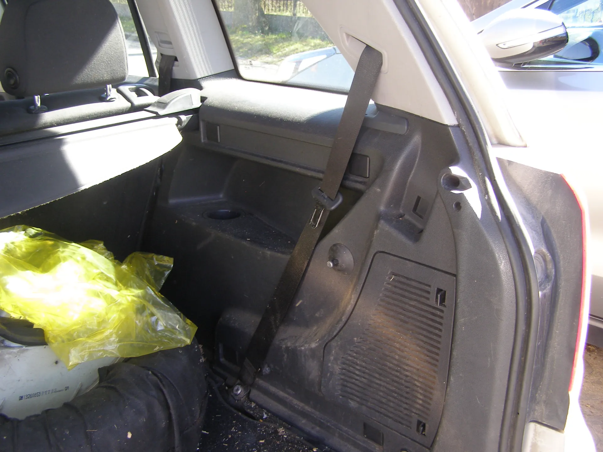 Wnętrze samochodu z widocznym tylnym siedzeniem, czarnym pasem bezpieczeństwa i żółtą plastikową torbą na lewej stronie. W tle przez otwarte drzwi widać fragment innego pojazdu oraz zieloną roślinność. Wnętrze posiada czarno-szare panele, uchwyt na napoje oraz siatkę wentylacyjną na drzwiach. Na podłodze widoczne są drobne zabrudzenia.