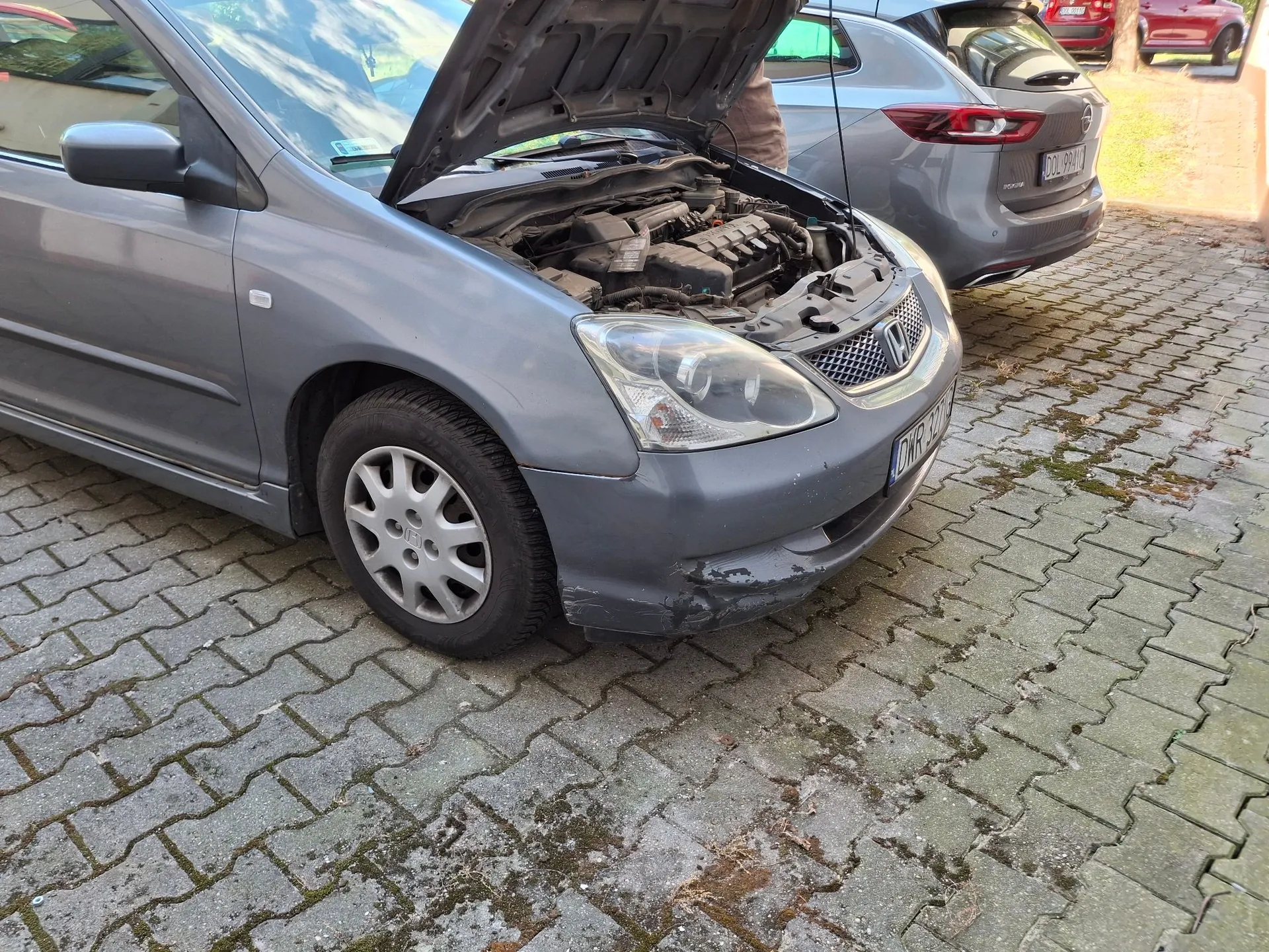Srebrny samochód osobowy marki Honda z otwartą maską, odsłaniającą silnik. Pojazd stoi na kostce brukowej. Na przednim zderzaku, po stronie pasażera, widoczne jest uszkodzenie w postaci zarysowań i odprysków lakieru. W tle widoczny jest inny, srebrny samochód.