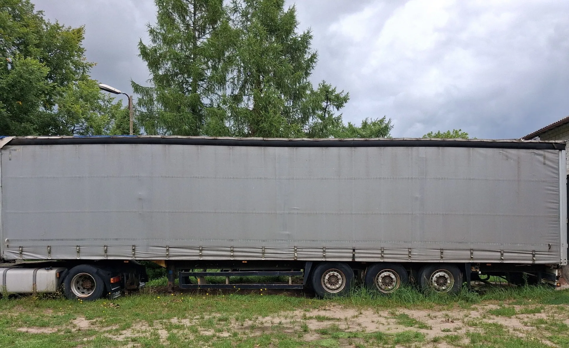 Boczny widok długiej, szarej naczepy ciężarowej z plandeką, zaparkowanej na trawiastym terenie. Naczepa posiada trzy osie z kołami po prawej stronie i jedną oś po lewej. W tle widoczne są zielone drzewa oraz zachmurzone niebo. Po lewej stronie znajduje się latarnia uliczna.