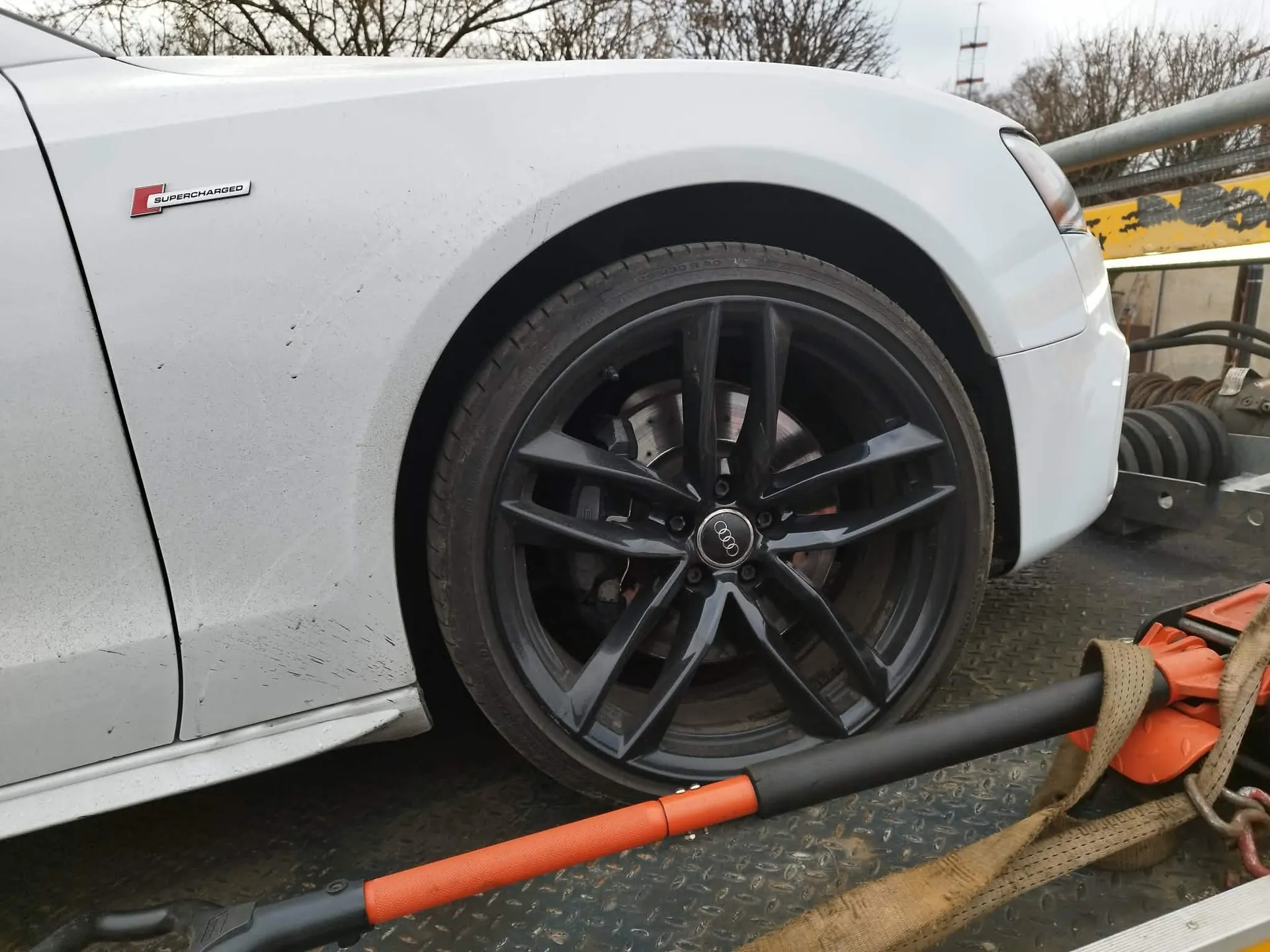 Zbliżenie na przednie prawe koło białego samochodu marki Audi, zaparkowanego na platformie lawety. Samochód posiada czarną felgę aluminiową z logo Audi w centrum. Na błotniku widoczny jest napis „SUPERCHARGED”. Pojazd jest zabezpieczony pomarańczowymi pasami transportowymi.