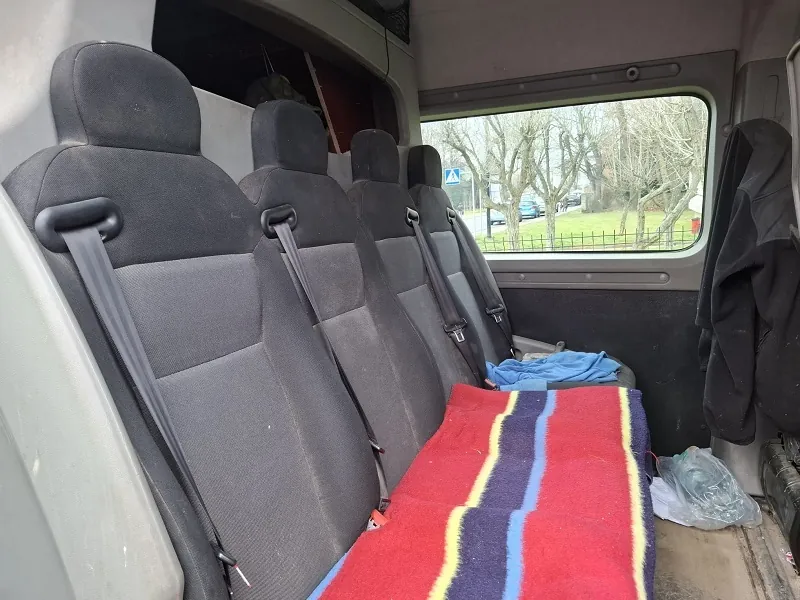 Wnętrze pojazdu z czterema szarymi fotelami pasażerskimi z pasami bezpieczeństwa. Przed fotelami leży czerwona narzuta z niebieskimi i żółtymi pionowymi pasami. Po prawej stronie widoczne są niebieska tkanina i przezroczysta torba plastikowa. Za oknem pojazdu widać drzewa, trawnik, ogrodzenie oraz niebieski znak drogowy w kształcie trójkąta. W tle droga z samochodami.