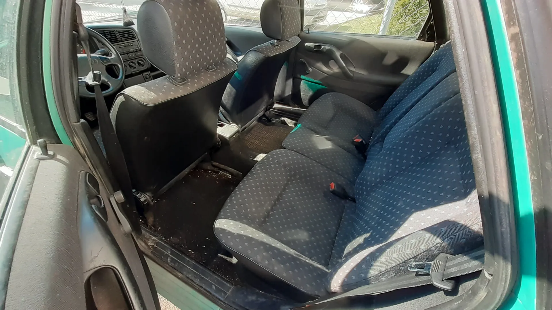 Wnętrze samochodu z otwartą lewą przodem drzwiami. Widoczne są czarne siedzenia z niebieskim wzorem kropek, kierownica po lewej stronie, deska rozdzielcza z przyciskami i wentylacją. Podłoga wewnątrz pojazdu jest zabrudzona. Zewnętrzna część drzwi i nadkola mają turkusowy kolor. W tle za samochodem widać siatkę ogrodzeniową oraz fragment innego pojazdu.