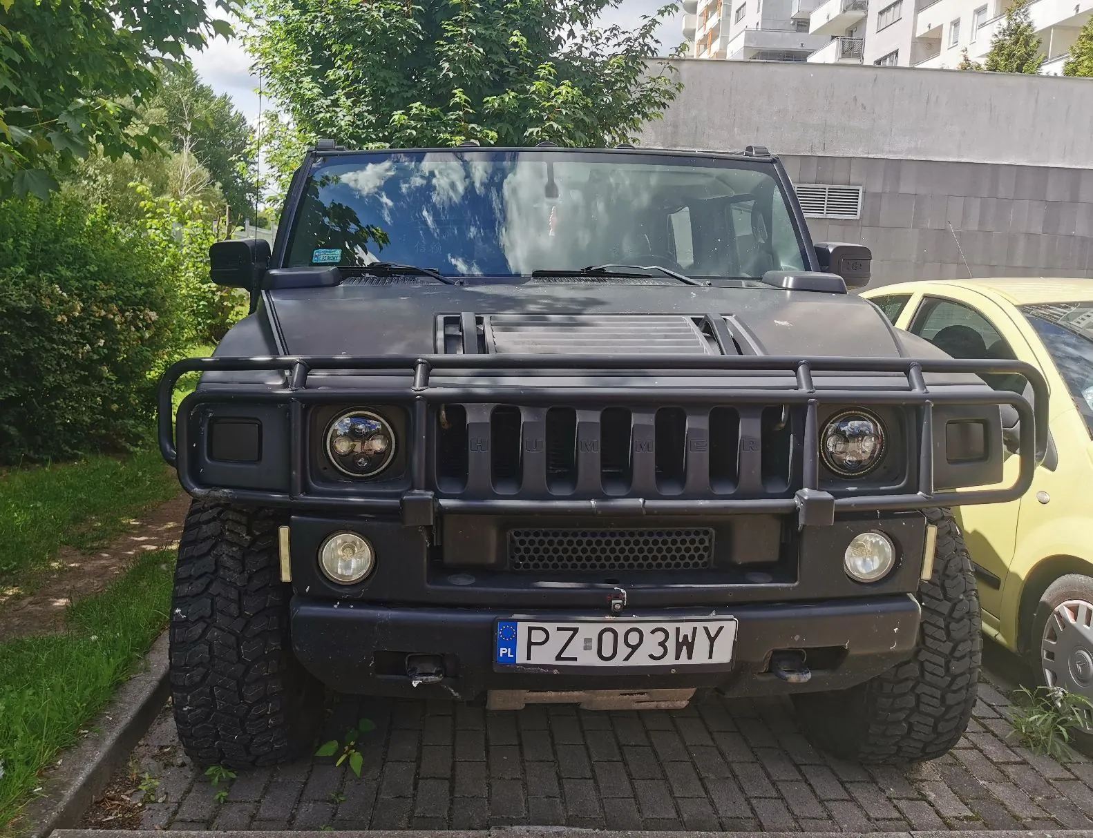 Czarny samochód terenowy marki Hummer zaparkowany przodem do obiektywu na kostce brukowej. Pojazd posiada masywny przedni zderzak z czarną rurą ochronną, duże terenowe opony oraz polską tablicę rejestracyjną o numerze PZ 093WY. W tle widoczne są zielone krzewy, drzewa oraz fragment jasnego budynku mieszkalnego. Po prawej stronie kadru widoczny jest fragment żółtego samochodu.