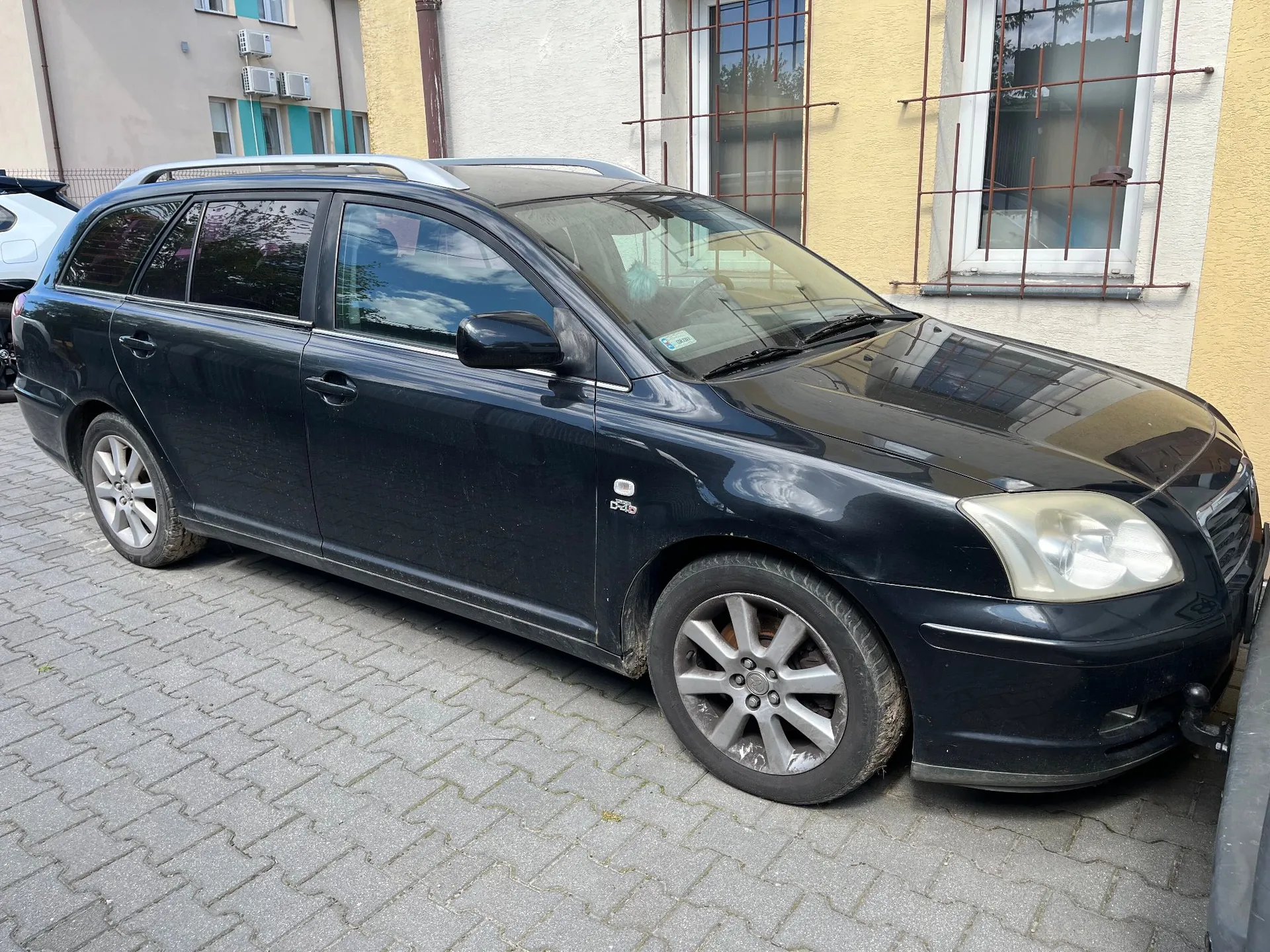 Czarny samochód osobowy typu kombi zaparkowany na kostce brukowej obok jasnej ściany budynku z oknami zabezpieczonymi kratami. Pojazd jest widoczny z profilu, z przodu i z boku.
