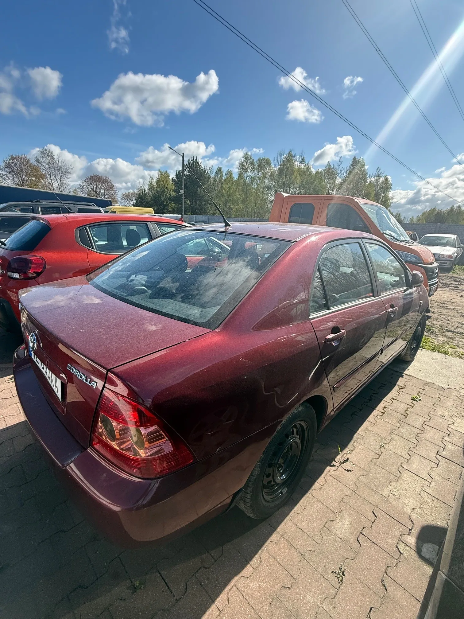 Bordowy samochód Toyota Corolla zaparkowany na brukowanej powierzchni. W tle widoczne są inne pojazdy, w tym czerwony samochód osobowy i pomarańczowy furgon. Na niebie białe chmury, drzewa oraz linie energetyczne. Na bagażniku widoczny napis 'COROLLA'.