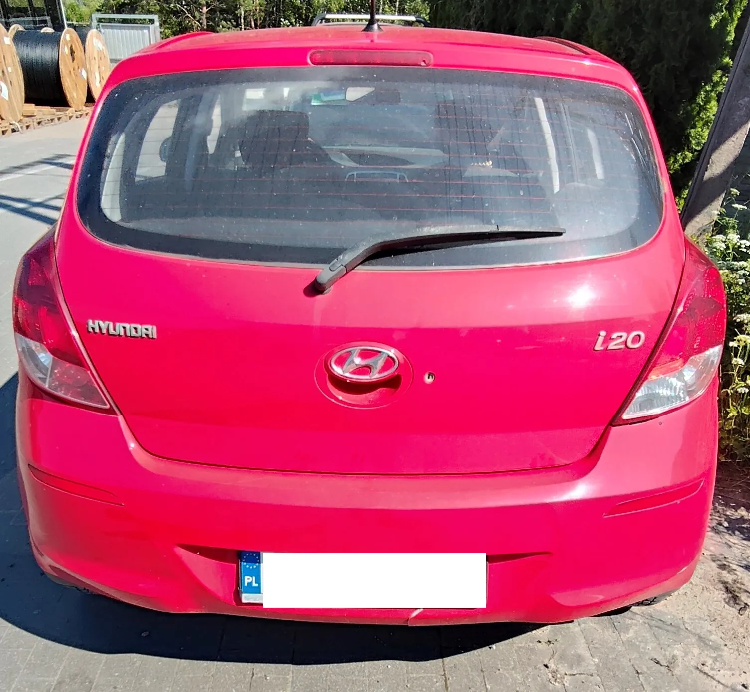Tylny widok czerwonego samochodu marki Hyundai i20. Na klapie bagażnika widoczne są napisy 'HYUNDAI' po lewej stronie, 'i20' po prawej oraz logo marki na środku. Samochód posiada tylną szybę z wycieraczką oraz tablicę rejestracyjną z oznaczeniem PL, która jest częściowo zakryta białym polem.