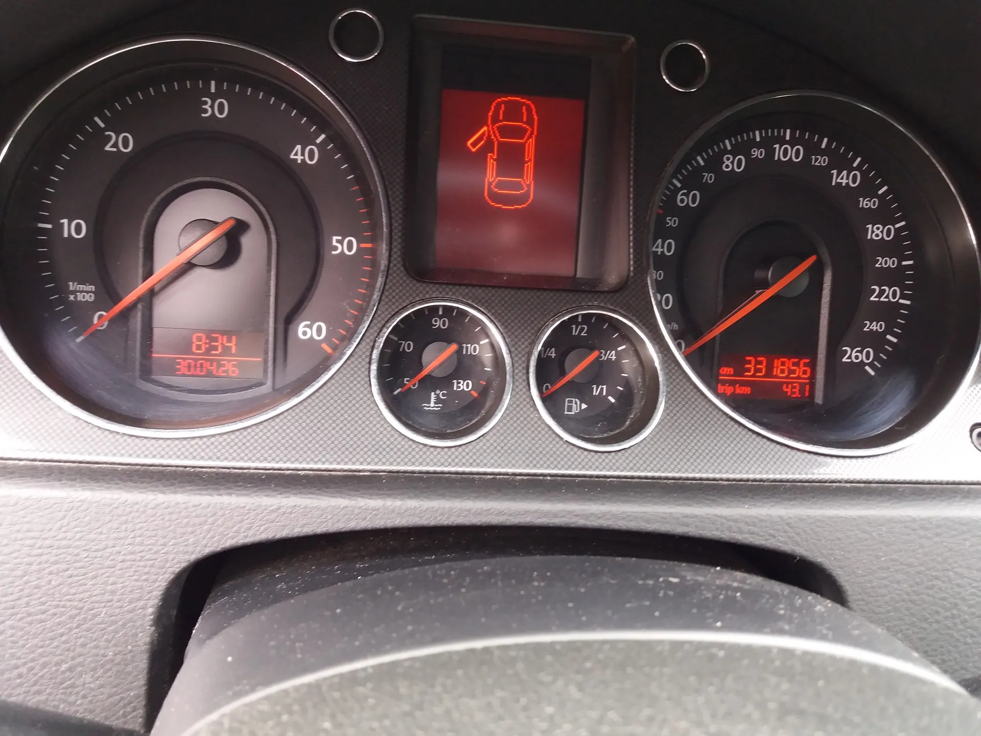 Tablica rozdzielcza samochodu z czarnym tłem i srebrnymi obramowaniami wskaźników. Po lewej stronie wskaźnik obrotomieru z zakresem 0-60 (1/min x100), czerwona igła wskazująca około 1. Poniżej cyfrowy wyświetlacz z czerwonymi cyframi: 8:34 i 30.04.26. W centrum górnej części czerwony symbol samochodu z otwartą klapą bagażnika na czerwonym tle. Poniżej dwa mniejsze wskaźniki: termometr (50-130°C) i wskaźnik paliwa (1/4 do 1). Po prawej stronie wskaźnik prędkości do 260 km/h, czerwona igła wskazująca około 40. Poniżej cyfrowy wyświetlacz z napisem "km 331856" i "trip km 43.1". Na dole widoczna część kierownicy. Wszystkie cyfry i symbole są czytelne, bez nachylenia.