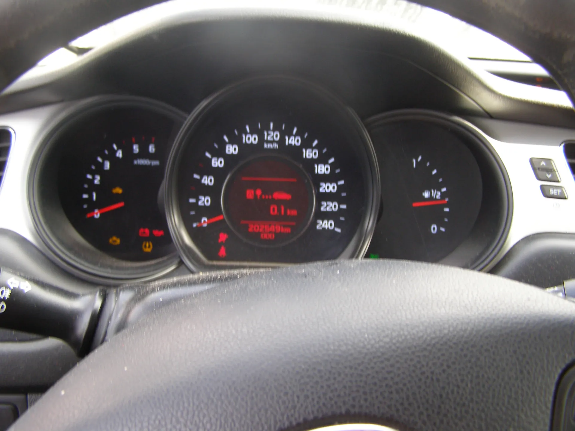 Zbliżenie na tablicę rozdzielczą samochodu z trzema głównymi wskaźnikami. W centrum znajduje się prędkościomierz z zakresem do 240 km/h, na wyświetlaczu cyfrowym widoczne wartości 0.1 km oraz 202549 km. Po lewej stronie tachometr z zakresem do 6 x1000 rpm, z czerwonymi i pomarańczowymi ikonami ostrzegawczymi. Po prawej wskaźnik paliwa z podziałką od 0 do 1. Na kierownicy widoczne przyciski sterujące. Wszystkie wskaźniki mają czarne tło z białymi i czerwonymi oznaczeniami.