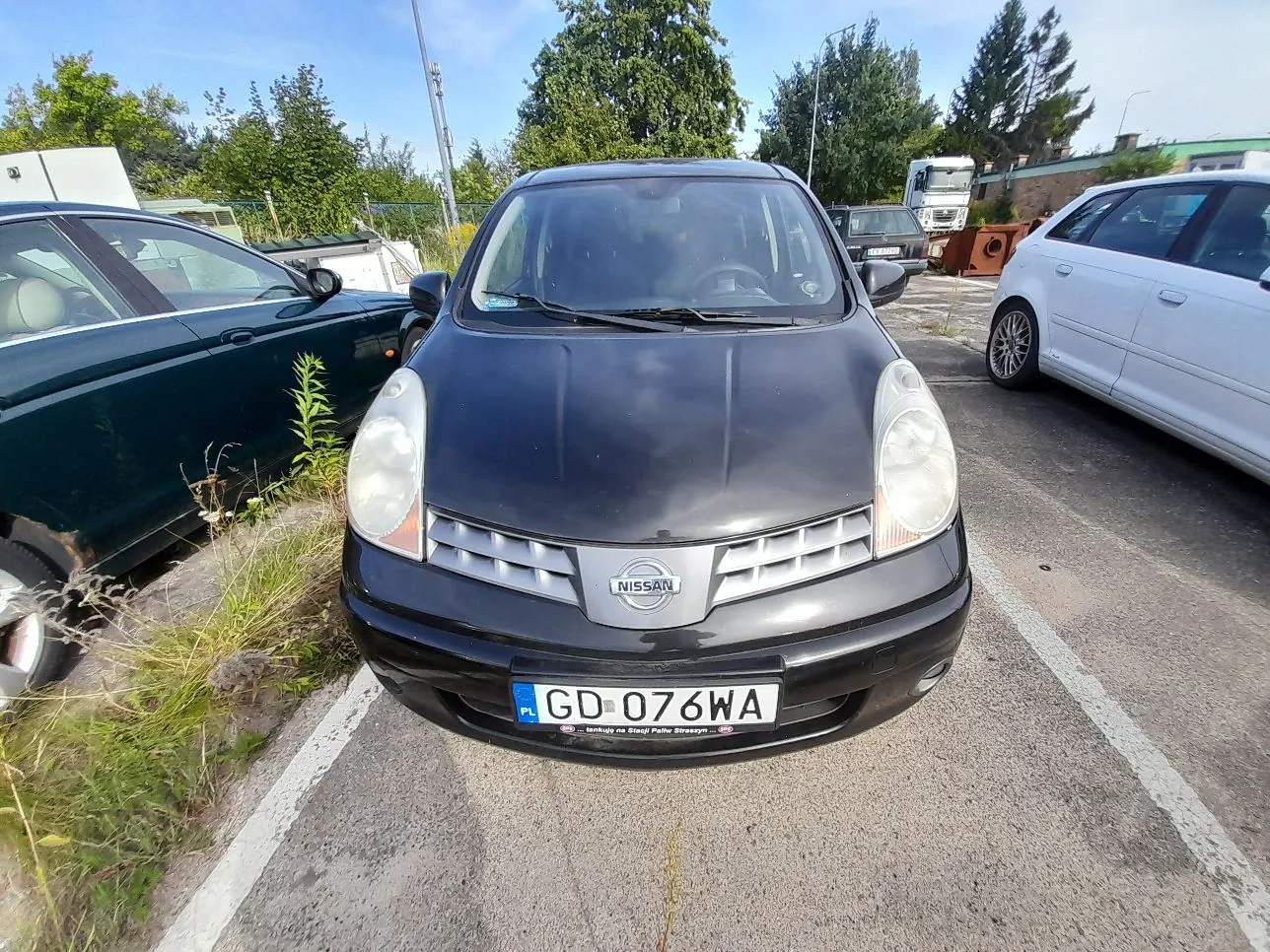 Czarny samochód marki Nissan zaparkowany na betonowym placu. Pojazd ma polską tablicę rejestracyjną o numerze GD 076WA. Po lewej stronie stoi ciemnozielony samochód, a po prawej srebrny. W tle widoczne są drzewa, niebo oraz biała ciężarówka.