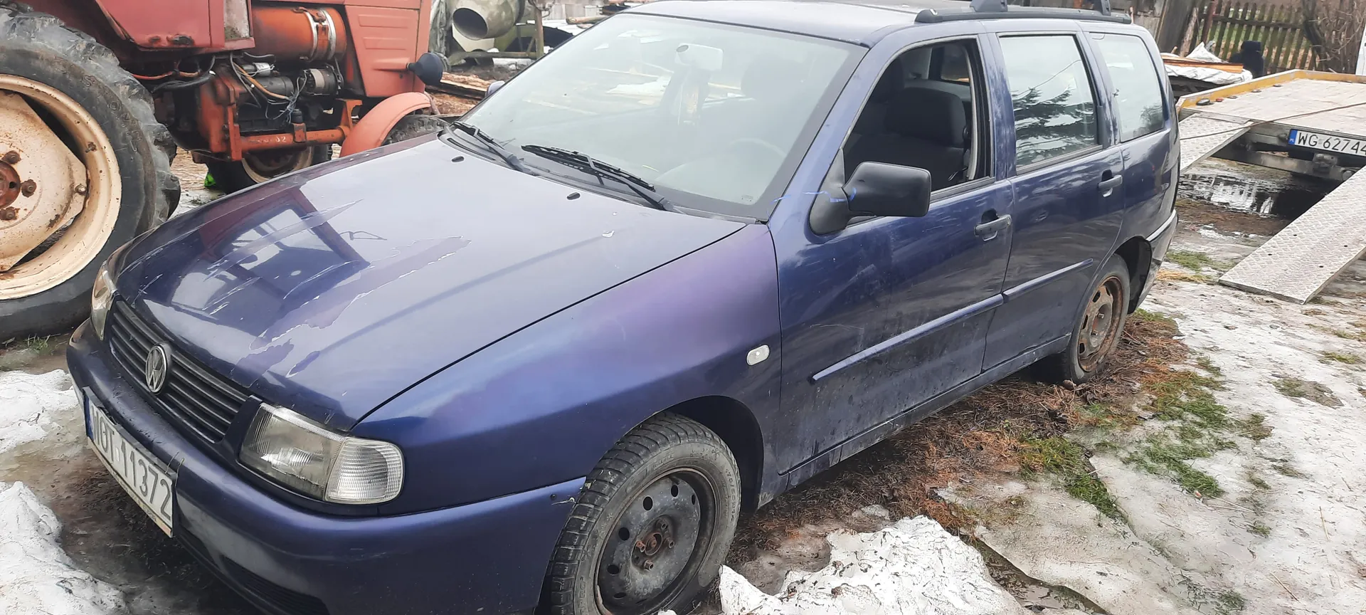 Niebieski samochód osobowy Volkswagen z tablicą rejestracyjną HOT 11372, z widocznymi uszkodzeniami i brudem, zaparkowany na śnieżno-błotnistym terenie. W tle czerwony traktor i przyczepa z tablicą WG 62744. Opony częściowo zużyte, otoczenie z zimowymi resztkami śniegu i trawą.