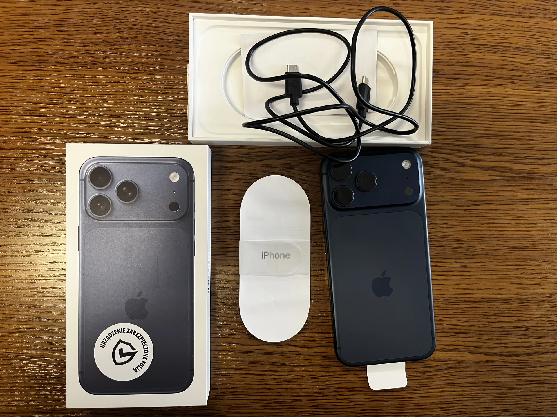 Zdjęcie przedstawia widok z góry na drewniany stół z kilkoma przedmiotami. Po lewej stronie znajduje się białe pudełko z czarnym iPhone'em, na którym widoczny jest okrągły naklejony znaczek z napisem "URZĄDZENIE ZABEZPIECZONE FOLIĄ" oraz logo. W centrum leży biały owalny przedmiot z napisem "iPhone". Po prawej stronie umieszczony jest czarny iPhone z widocznym logo Apple na tylnej klapce. Powyżej, w białym pudełku, znajduje się czarny kabel USB-C zakończony dwoma wtyczkami. Wszystkie elementy są ułożone równolegle do krawędzi zdjęcia.
