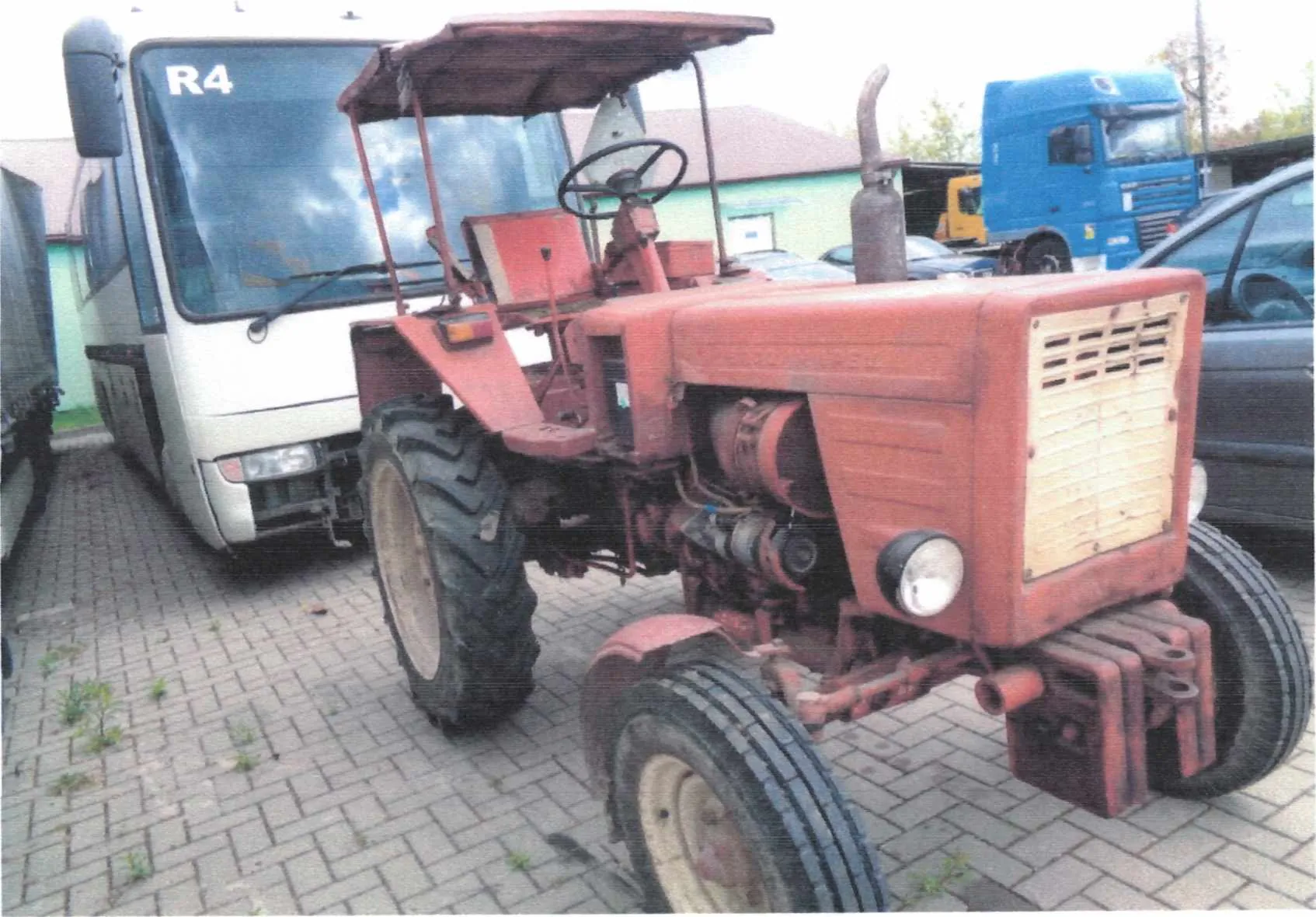 Czerwony traktor z daszkiem, kierownicą i dużymi oponami, zaparkowany na brukowanym placu. W tle biały autobus z napisem R4 na przedniej szybie, niebieska ciężarówka, czarny samochód osobowy oraz budynki z zielonymi i czerwonymi dachami. Na traktorze widoczne są reflektory, wydech i beżowa panelowa osłona z otworami wentylacyjnymi.