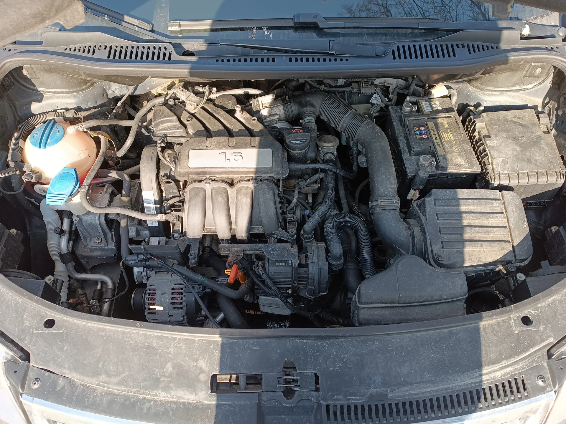 Widok otwartej tylnej części silnika samochodu z widocznym blokiem oznaczonym '1.6'. Po lewej stronie znajduje się zbiornik chłodniczy z niebieskimi korkami, a po prawej bateria z etykietą zawierającą kolorowe symbole. W centralnej części widoczne są czarne przewody, rury wentylacyjne oraz elementy układu elektrycznego. Na dole kadru widoczna jest część maski z otworami wentylacyjnymi. Całość pokryta jest warstwą kurzu.