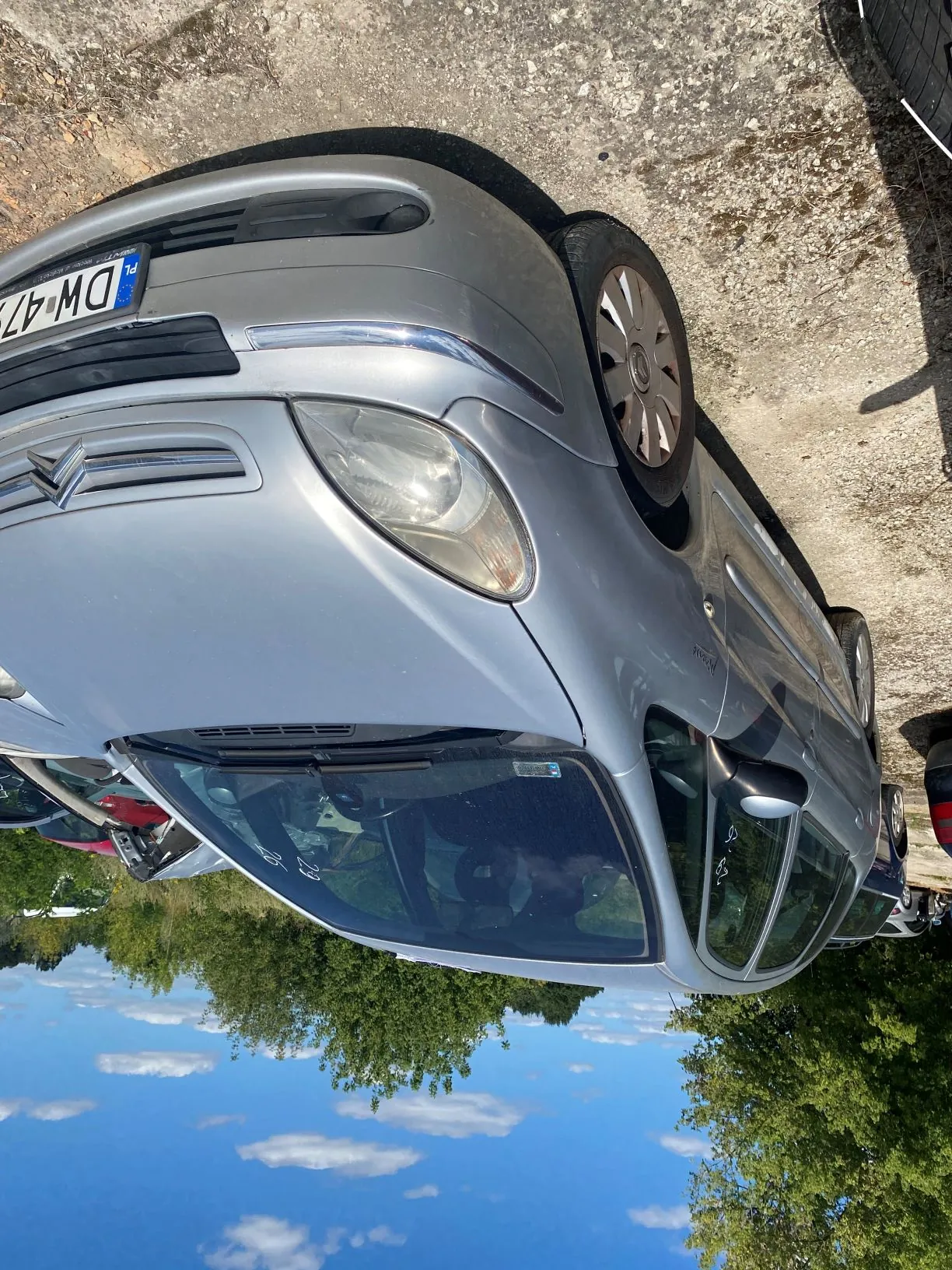 Srebrny samochód marki Citroën zaparkowany na piaszczystym podłożu. Widoczny jest przód pojazdu z fragmentem tablicy rejestracyjnej o numerze DW 47. W tle znajdują się zielone drzewa oraz niebieskie niebo z białymi chmurami.