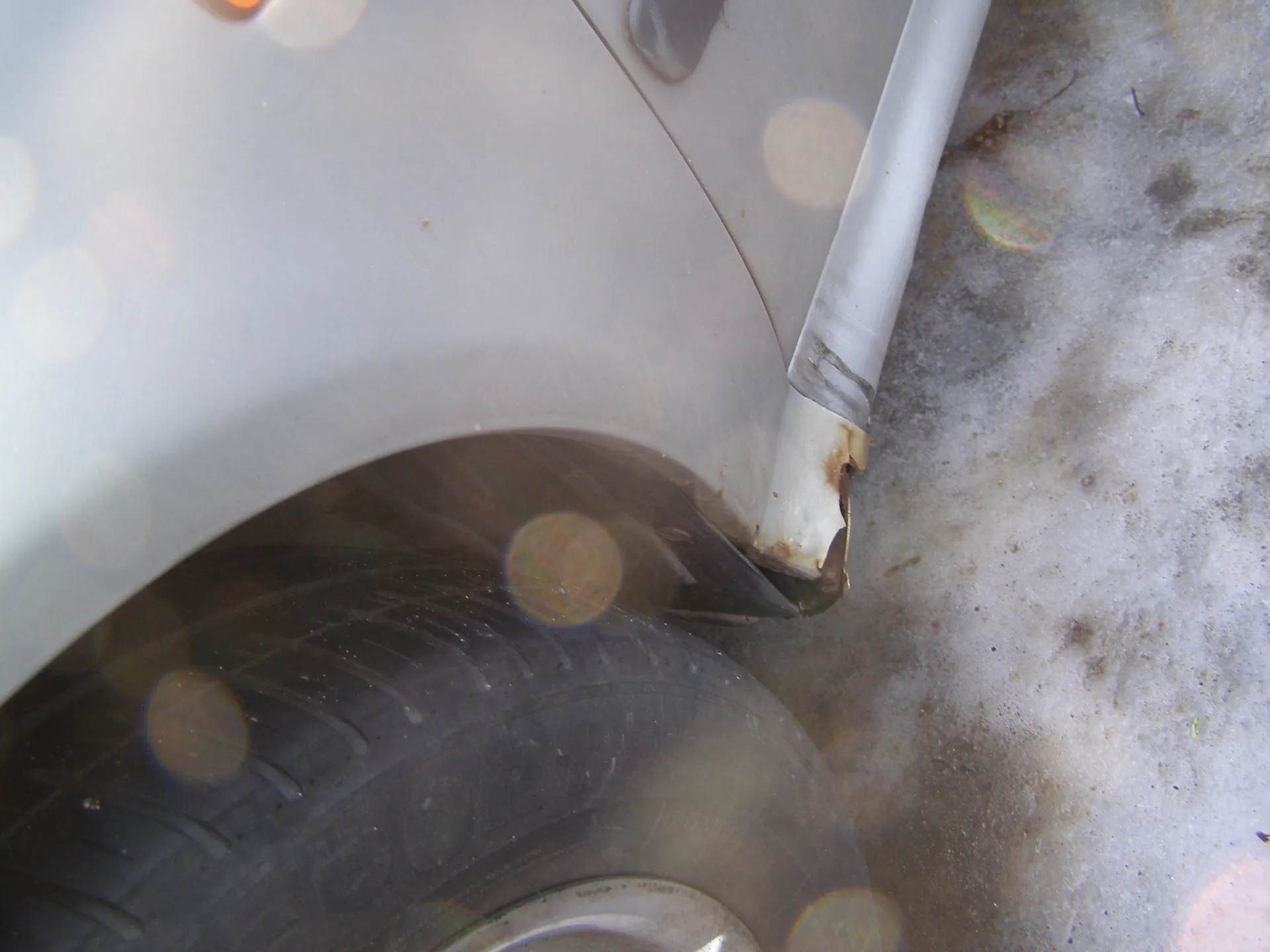 Zbliżenie na fragment srebrnego samochodu, widoczna opona z częściowo czytelnym napisem, uszkodzona biała obudowa kołowa z oznakami korozji, tło pokryte śniegiem lub lodem z plamami brudu. Na obrazie widoczne są efekty świetlne w postaci rozmytych okręgów.