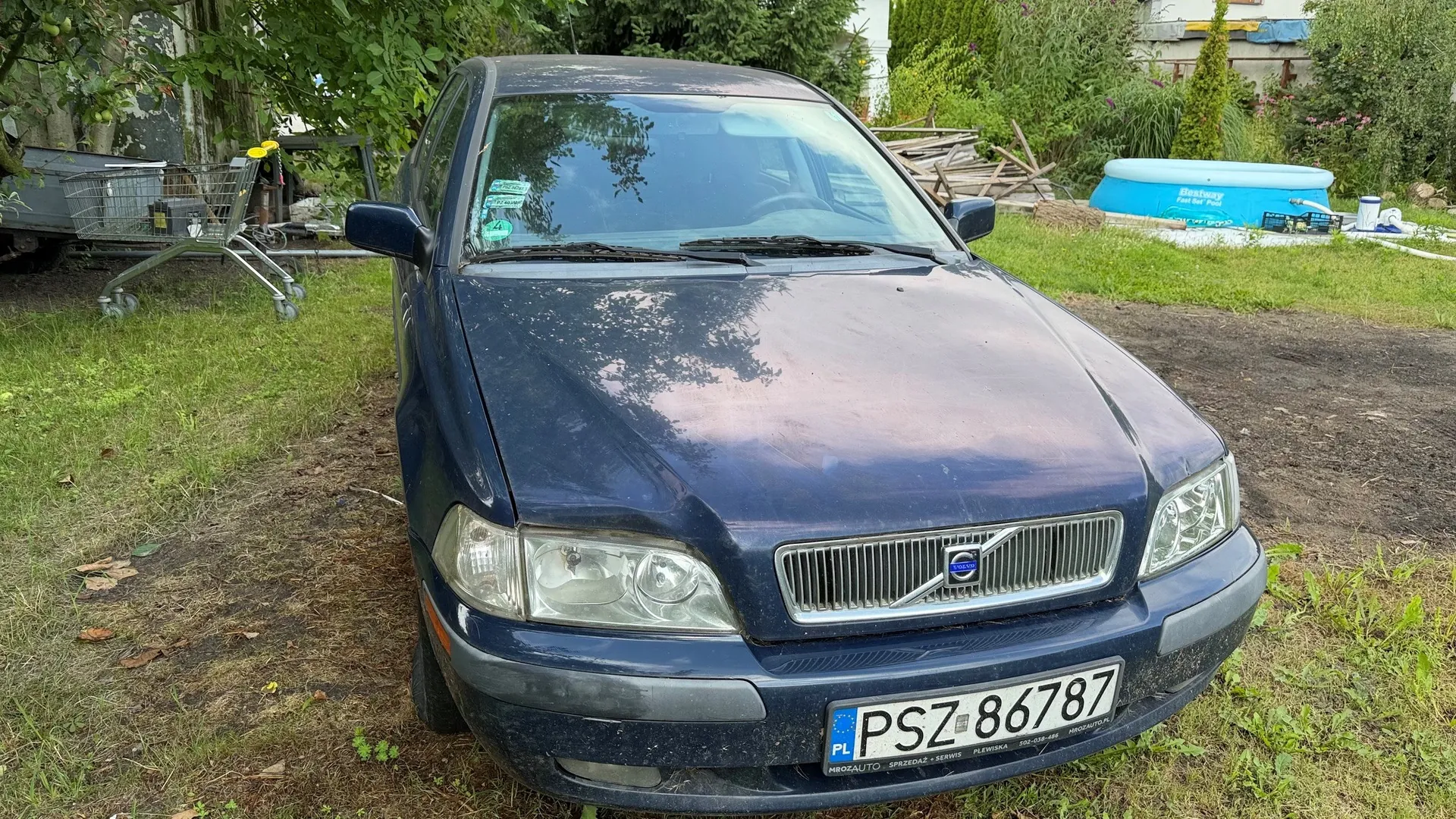 Granatowy samochód marki Volvo zaparkowany na trawie. Widoczny jest przód pojazdu z grillem, logo Volvo oraz polską tablicą rejestracyjną o numerze PSZ 86787. W tle po lewej stronie znajduje się metalowy wózek sklepowy, a po prawej niebieski basen ogrodowy marki Bestway.