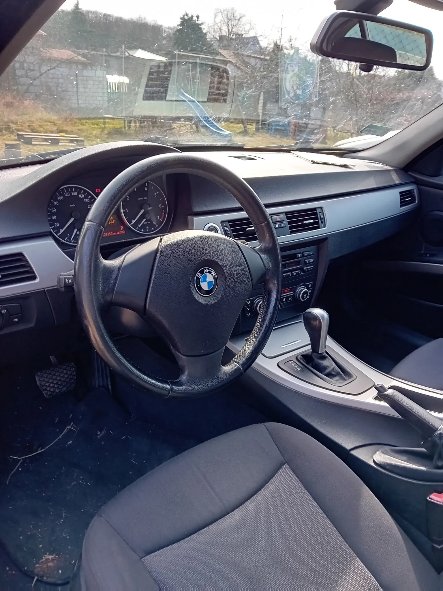 Wnętrze samochodu marki BMW z widocznym kierownicą z logo firmy, tablicą rozdzielczą z prędkościomierzem i tachometrem, centralną konsolą z systemem audio i klimatyzacją oraz dźwignią zmiany biegów. Przednie siedzenia są szare, a na podłodze widoczne są drobne śmieci. Przez przednią szybę widać budynek, drzewa i niebieską poślizgówkę na zewnątrz. Lusterko wsteczne umieszczone jest na górze zdjęcia.