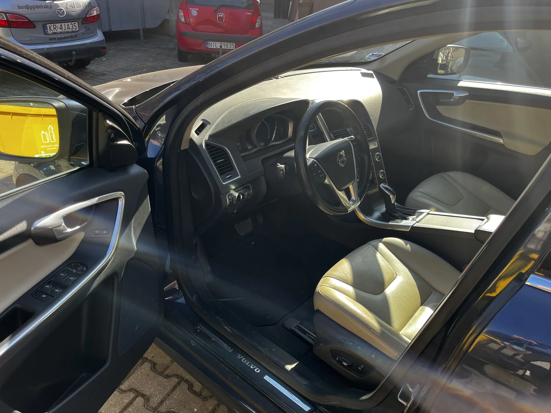 Wnętrze czarnego samochodu marki Volvo z otwartą przodem drzwiami kierowcy. Widoczne są beżowe fotele, kierownica z logo Volvo, deska rozdzielcza, przyciski sterujące i automatyczna skrzynia biegów. Na progu drzwi widnieje napis 'VOLVO'. Poza samochodem, na tle brukowanej nawierzchni, stoją dwa inne pojazdy: srebrny Mazda z tablicą rejestracyjną KR-4JA35 oraz czerwony samochód z tablicą N1L 71835. W lusterku bocznym głównego samochodu widoczny jest żółty naklejony znak z symbolami butelek.