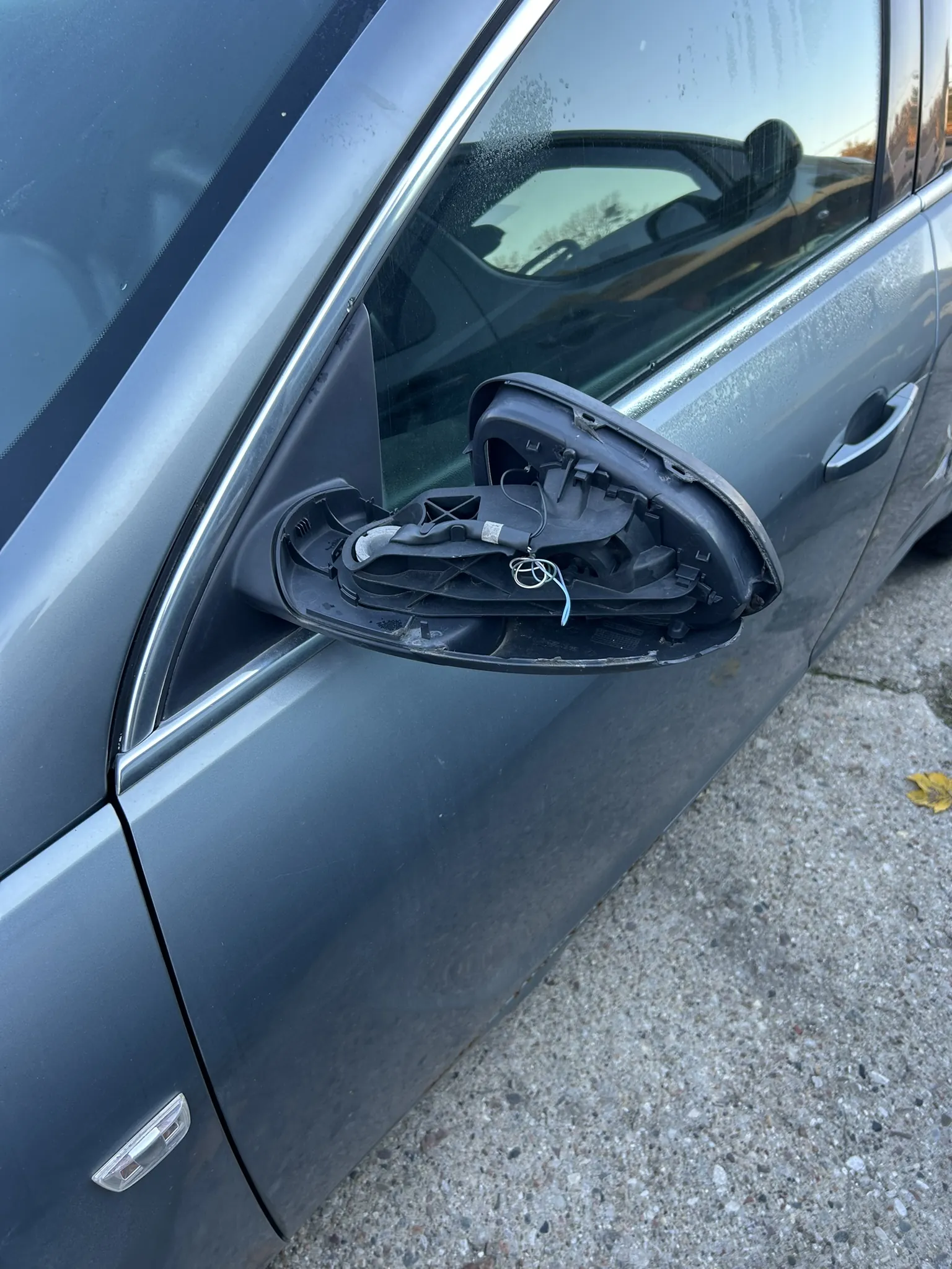 Zdjęcie przedstawia szary samochód z uszkodzonym lusterkiem bocznym. Obudowa lusterka jest otwarta, odsłaniając wewnętrzne elementy i przewody. Samochód zaparkowany jest na betonowej powierzchni, na której widoczne są liście. W szybie odbija się inny pojazd oraz drzewa.