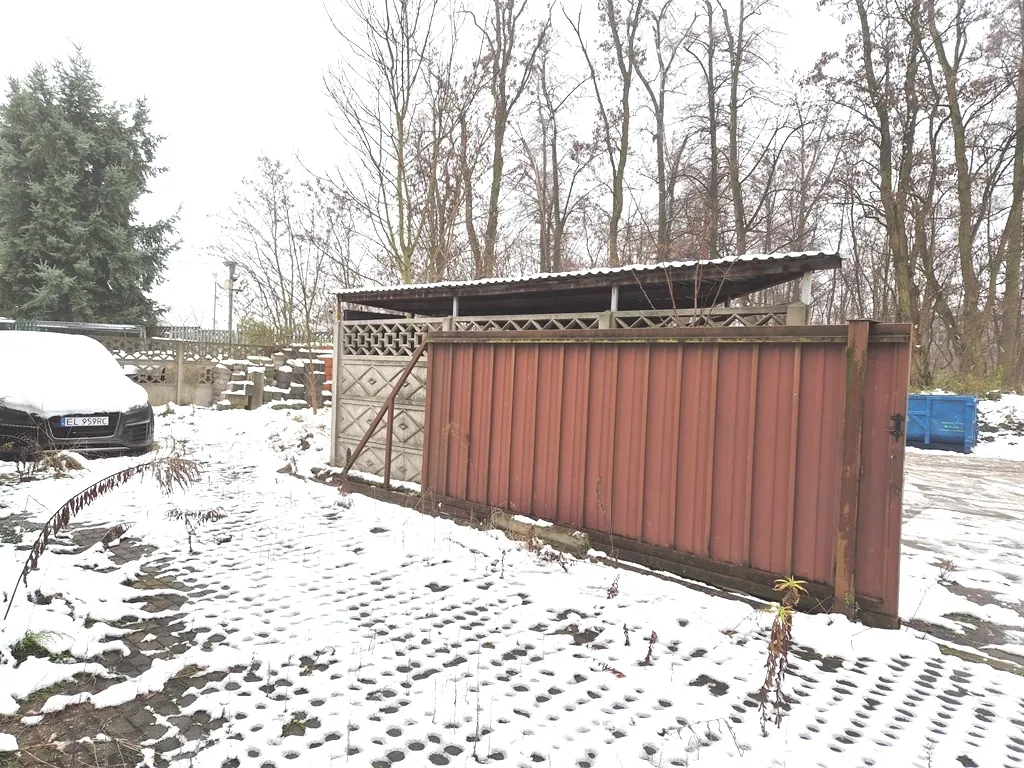 Zdjęcie przedstawia podwórze pokryte cienką warstwą śniegu. Po lewej stronie widoczny jest fragment białego samochodu z tablicą rejestracyjną EL 959RC. W centrum znajduje się drewniana, brązowa konstrukcja przypominająca wiatę lub ogrodzenie z daszkiem. W tle widać bezlistne drzewa i niebo w kolorze jasnoszarym. Po prawej stronie znajduje się niebieski kontener na odpady.