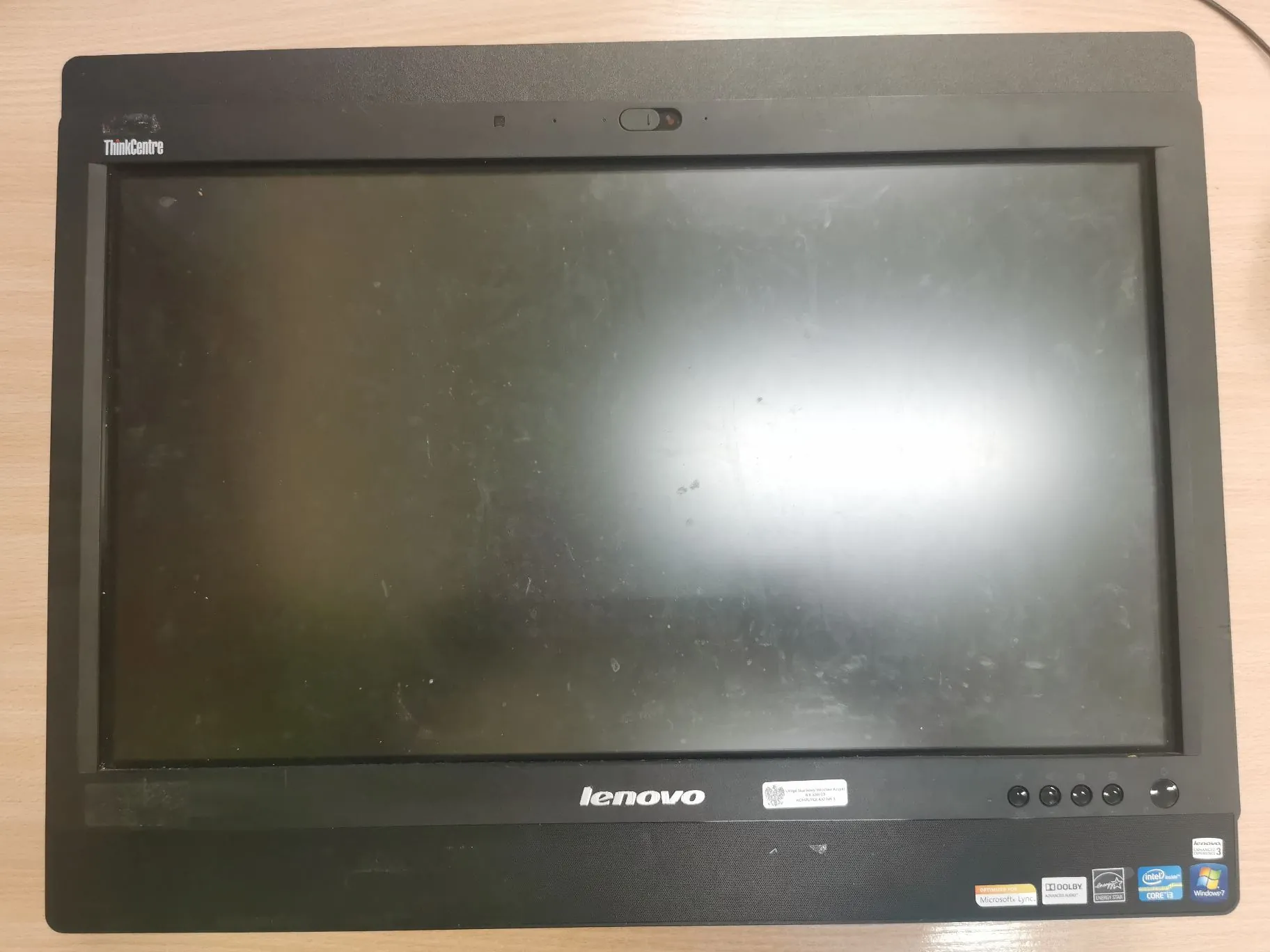 Czarny monitor marki Lenovo ThinkCentre z zakurzonym ekranem. Na górnej ramce widnieje napis 'ThinkCentre', a na dolnej logo 'lenovo'. W prawym dolnym rogu znajdują się naklejki z informacjami o sprzęcie, w tym logo Intel Core i5 oraz Windows 7. Obok naklejek znajdują się przyciski sterujące.