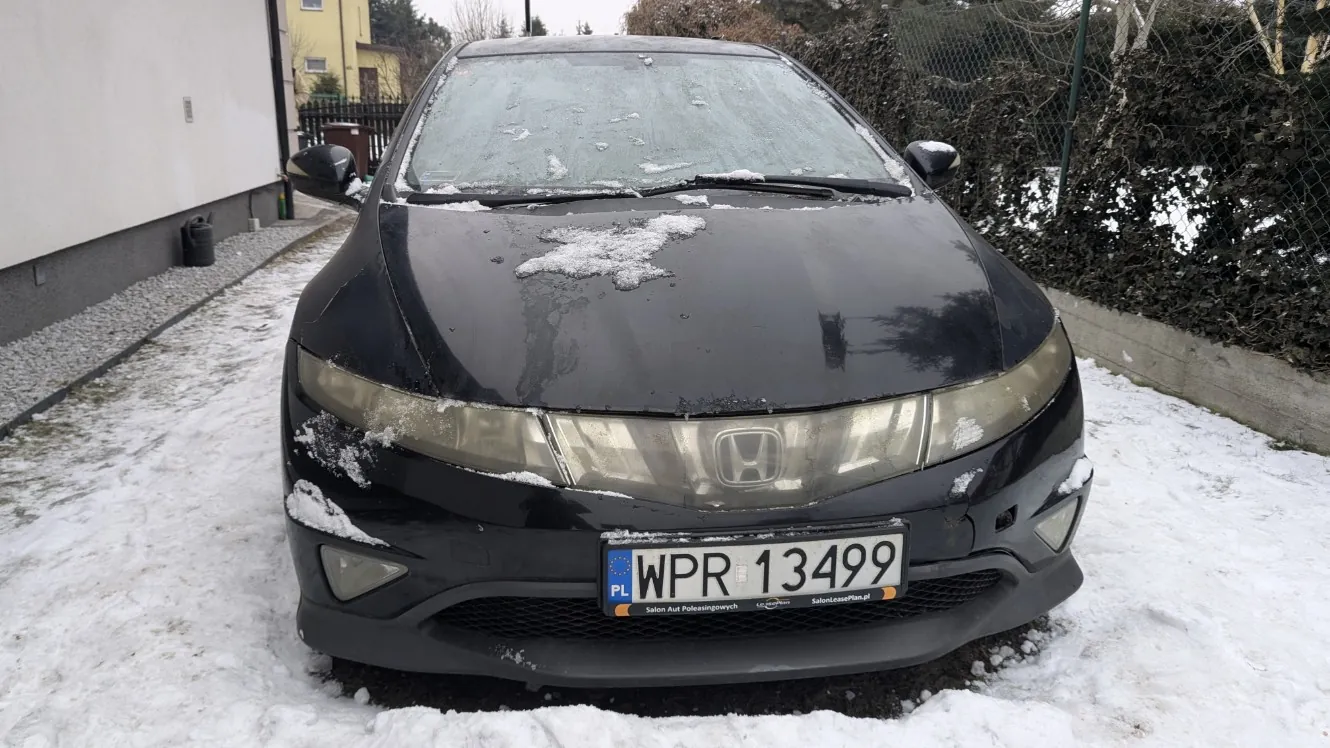 Czarny samochód marki Honda zaparkowany na śnieżnej powierzchni. Na masce i przedniej szybie widoczne są resztki śniegu. Tablica rejestracyjna z białym tłem i czarnym napisem WPR 13499, z lewej strony niebieski pas z napisem PL oraz dodatkowymi napisami Salon Aut Polazkowych i SalonSamochodowy.pl. W tle budynek z białą ścianą po lewej stronie, ogrodzenie z siatki i krzewy po prawej, a także domy i drzewa w oddali. Podłoga pokryta jest śniegiem i żwirem.