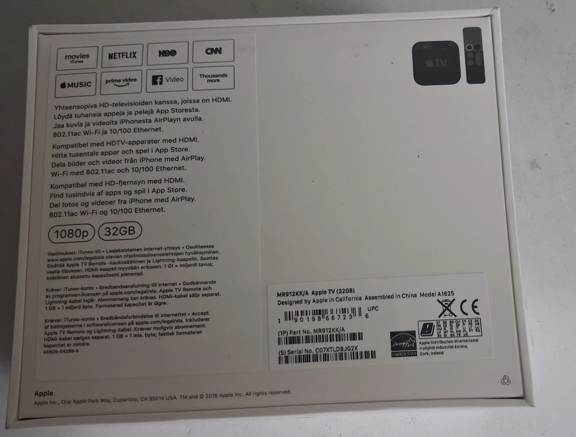Zdjęcie tylnej części białego opakowania Apple TV (32GB). W górnej części znajdują się ikony aplikacji: movies (iTunes), NETFLIX, HBO, CNN, MUSIC, prime video, f Video oraz napis 'Thousands more'. Poniżej znajduje się opis produktu w językach fińskim, szwedzkim i norweskim. Widoczna jest informacja o rozdzielczości '1080p 32GB'. W dolnej części znajdują się kody kreskowe, numery seryjne, oznaczenia certyfikacji (CE, XC), informacja 'Designed by Apple in California Assembled in China Model A1625' oraz dane adresowe firmy Apple Inc. w Cupertino, USA.
