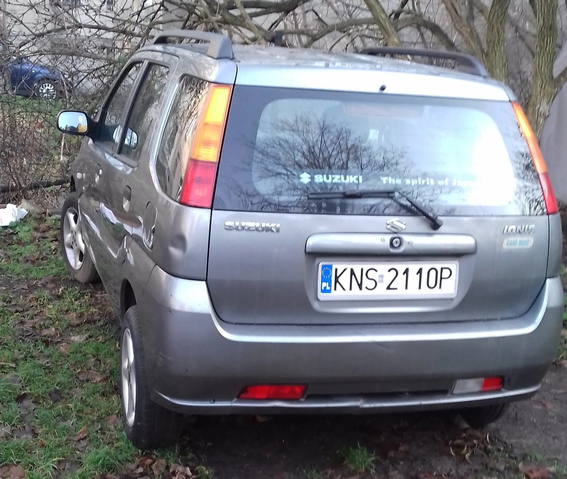 Srebrny samochód marki Suzuki Ignis zaparkowany na trawie. Widok z tyłu i z boku. Na tylnej szybie znajduje się napis 'SUZUKI The spirit of Japan'. Samochód posiada polską tablicę rejestracyjną o numerze KNS 2110P.