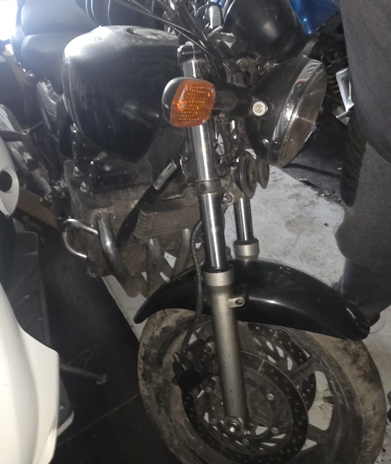 Zbliżenie na przednią część czarnego motocykla, w tym reflektor, pomarańczowy kierunkowskaz, przednie koło z dyskiem hamulcowym oraz fragment silnika. Widoczne są również elementy otoczenia: betonowa podłoga, fragment białego pojazdu po lewej stronie oraz noga osoby w ciemnych spodniach po prawej. Motocykl wydaje się nieco zabrudzony.