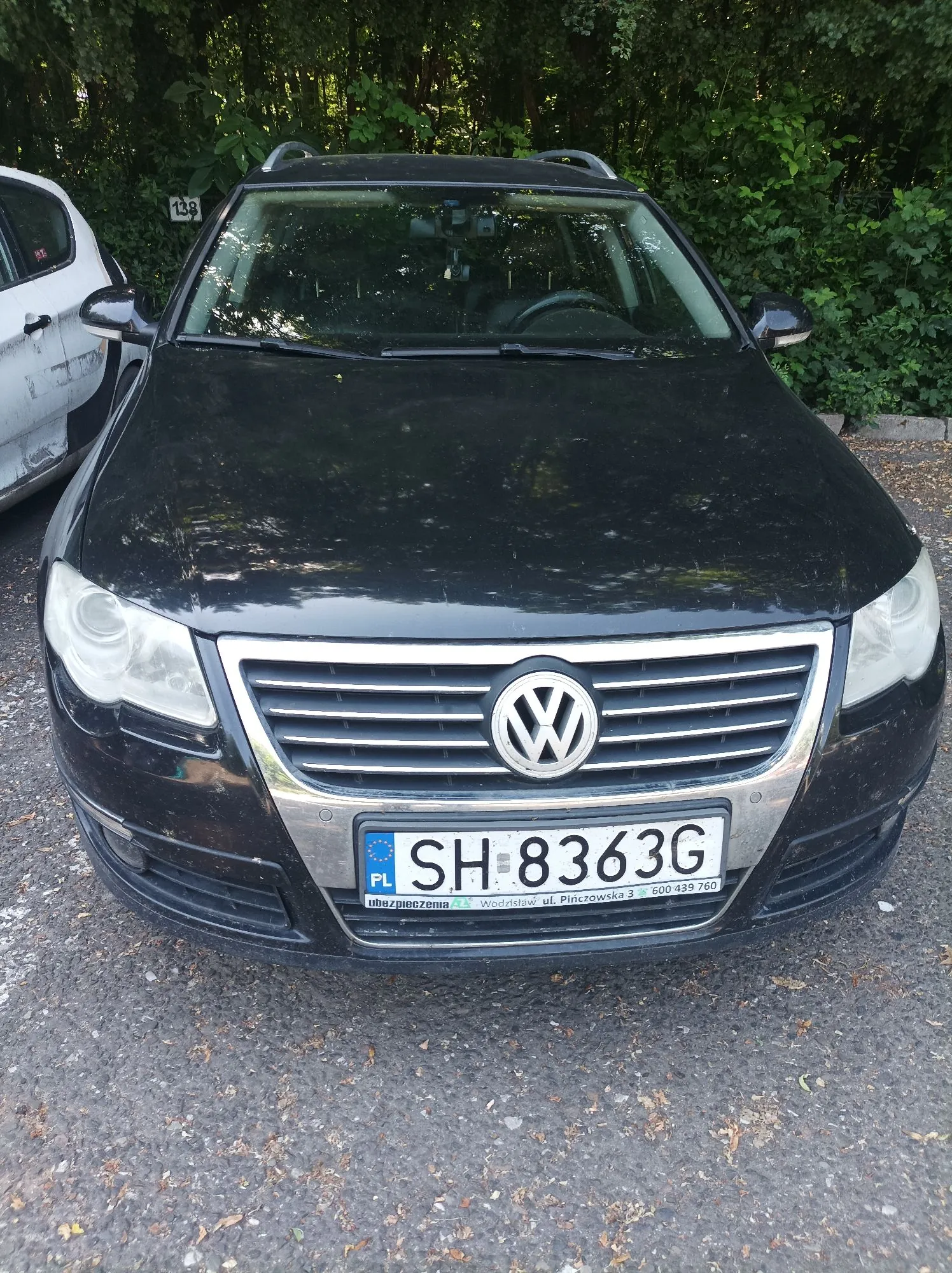 Czarny samochód marki Volkswagen zaparkowany na szutrowym podłożu na tle zielonych krzewów. Widoczny jest przód pojazdu z grillem, logo producenta oraz polską tablicą rejestracyjną o numerze SH 8363G. Poniżej tablicy znajduje się ramka z napisem 'ubezpieczenia.pl Włodzimierz ul. Piłcrowska 3 800 439 760'. Po lewej stronie widoczny jest fragment białego samochodu.