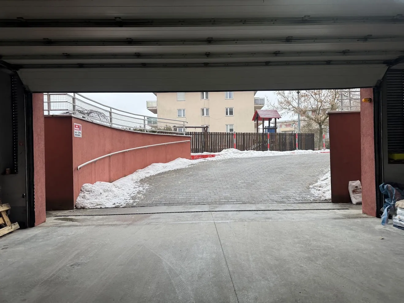 Widok z wnętrza garażu lub hali na zewnątrz. W kadrze znajduje się betonowy podjazd prowadzący w górę, po bokach którego leżą hałdy białego śniegu. Po lewej stronie widoczny jest czerwony mur z białą poręczą. W tle znajduje się brązowy płot, wielopiętrowy budynek mieszkalny w kolorze beżowym oraz mały drewniany domek z czerwonym dachem. Niebo jest zachmurzone.