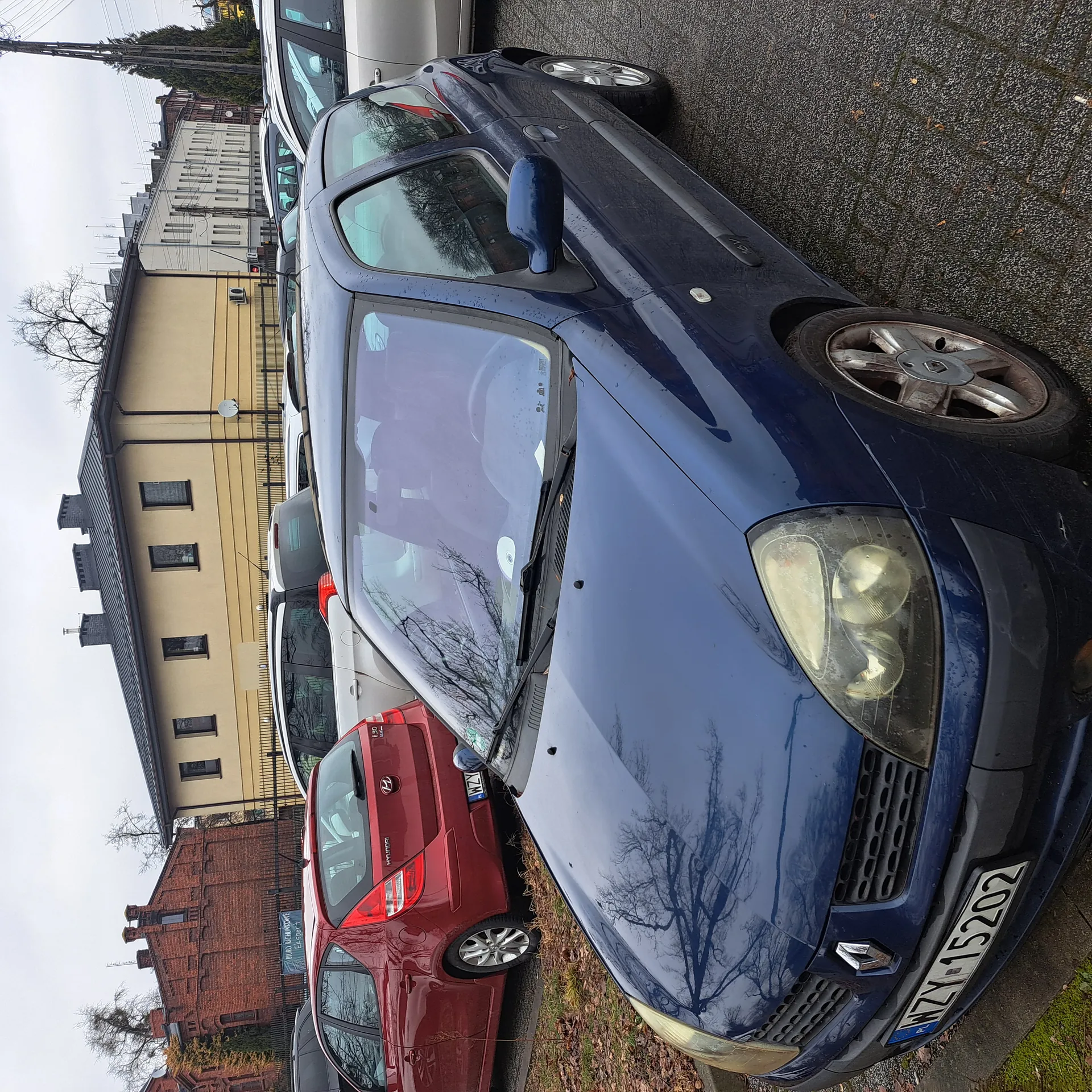 Zdjęcie przedstawia parking z kilkoma samochodami. W pierwszym planie niebieski samochód marki Renault z tablicą rejestracyjną WZY 15202. Za nim widoczne są inne pojazdy, w tym czerwony Hyundai i srebrny samochód. W tle stoją budynki: żółty dom z czarnym dachem, ceglany budynek oraz biały blok. Na niebie panuje pochmurna pogoda. Na ziemi leżą suche liście, a asfaltowy parking ma drobne nierówności.