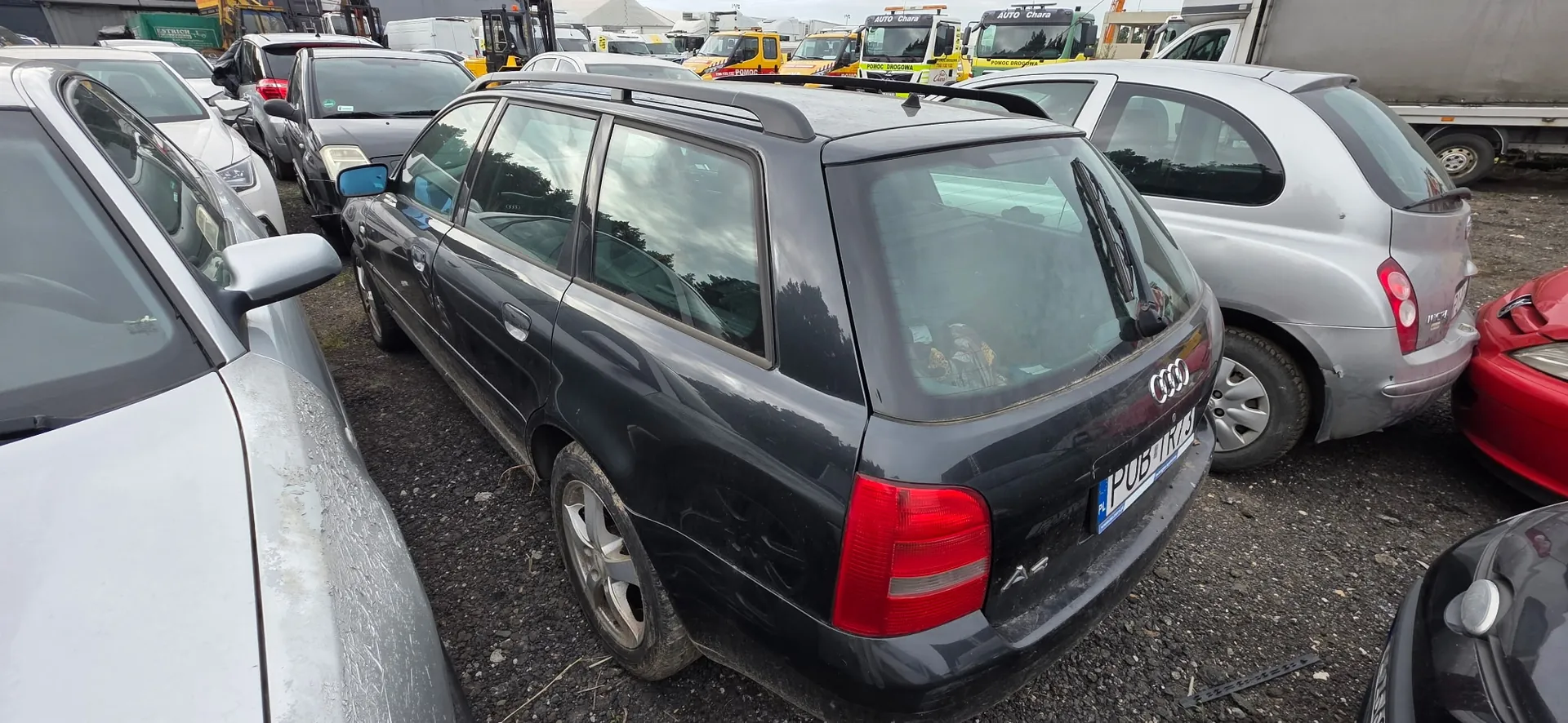 Zdjęcie przedstawiające zaparkowane samochody na placu. Na pierwszym planie znajduje się ciemnoszary samochód marki Audi w nadwoziu kombi, widziany od tyłu i z boku. Samochód ma tablicę rejestracyjną o numerze PUB 176Z. Po lewej stronie widoczny jest fragment srebrnego samochodu, a po prawej srebrny samochód kompaktowy oraz fragment czerwonego pojazdu. W tle widać inne samochody oraz żółte pojazdy specjalistyczne.