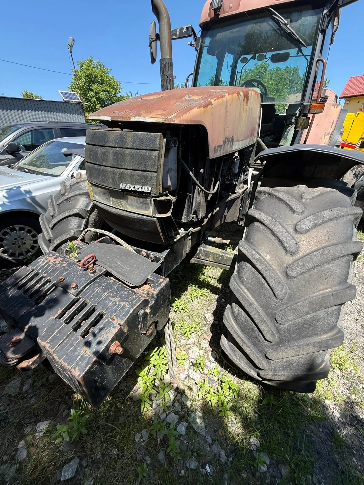 Zbliżenie na przód starego, zardzewiałego traktora marki Maxxam. Maszyna ma duże, błotniste opony z głębokim bieżnikiem oraz ciężki, czarny obciążnik zamontowany z przodu. W tle widoczne są zaparkowane samochody, budynki oraz niebo.