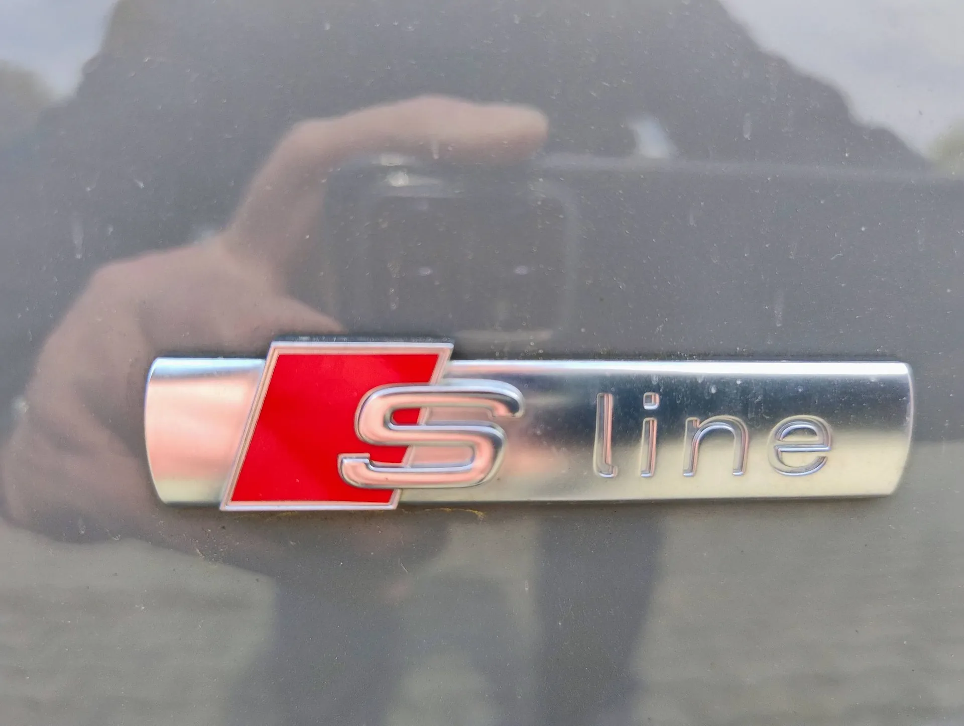 Zbliżenie na srebrny emblemat z napisem „S line” na karoserii samochodu. Litera „S” znajduje się na czerwonym tle w kształcie równoległoboku, a słowo „line” jest napisane obok srebrnymi, wypukłymi literami. W lakierze samochodu widoczne jest odbicie osoby robiącej zdjęcie.