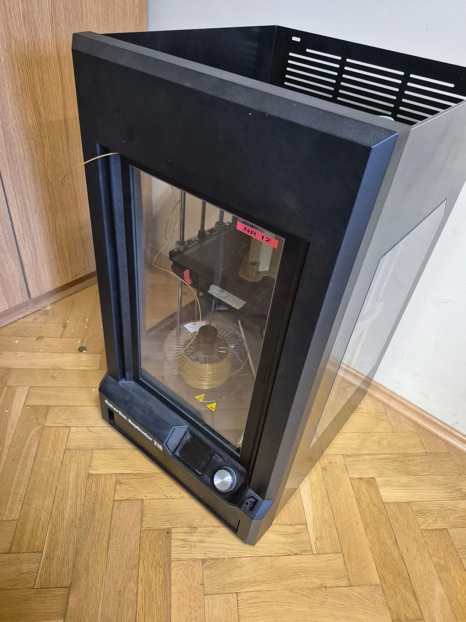 Czarna drukarka 3D marki MakerBot Replicator 2 Pro w zamkniętej obudowie z przezroczystą szybą z przodu i z boku. Wewnątrz urządzenia widoczna jest szpula z filamentem oraz mechanizm drukujący. Na obudowie znajduje się czerwona naklejka z napisem 'NR-12' oraz żółte trójkątne znaki ostrzegawcze. Na dolnej krawędzi widnieje napis 'MakerBot Replicator 2 Pro'. Urządzenie stoi na drewnianej podłodze ułożonej w jodełkę, obok jasnej ściany i drewnianych mebli.