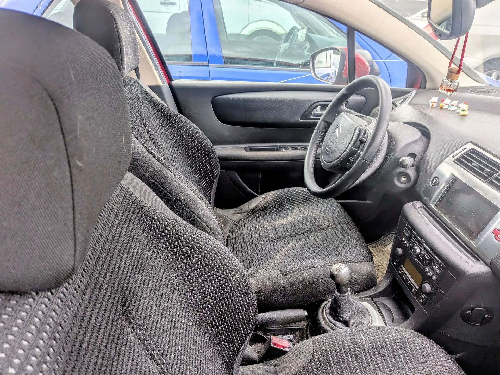 Wnętrze samochodu z perspektywy tylnego siedzenia, ukazujące przednie fotele z czarnej, teksturowanej tkaniny. Widoczna jest kierownica z logo Citroëna, deska rozdzielcza z radiem i ekranem, oraz lewarek zmiany biegów. Przez przednią szybę i okno widać fragmenty innych samochodów w kolorach niebieskim i czerwonym.