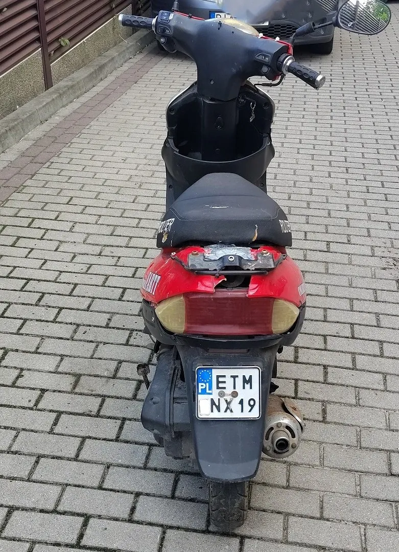 Widok z tyłu na czerwono-czarny skuter zaparkowany na kostce brukowej. Pojazd jest uszkodzony w tylnej części nadkoli i pod siedzeniem. Skuter posiada polską tablicę rejestracyjną o numerze ETM NX19.