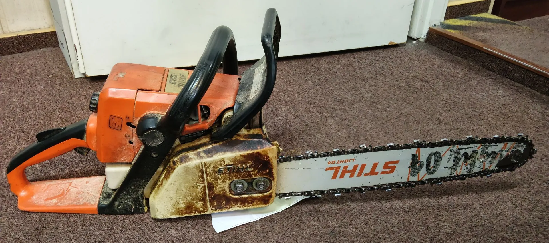 Pomarańczowa piła spalinowa marki STIHL leżąca poziomo na szarym dywanie. Urządzenie jest zabrudzone, a na prowadnicy łańcucha widnieje napis 'STIHL' oraz oznaczenie 'LIGHT 04'.