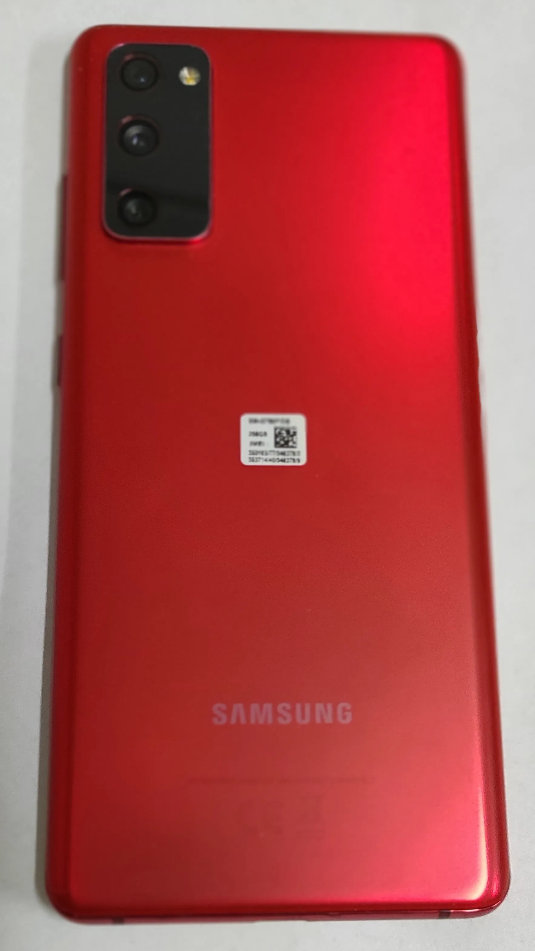 Tylna obudowa czerwonego smartfona marki Samsung. W lewym górnym rogu znajduje się prostokątny moduł z trzema obiektywami aparatu i lampą błyskową. Na środku obudowy naklejona jest mała biała etykieta z kodem QR i tekstem. Na dole znajduje się srebrny napis 'SAMSUNG'.