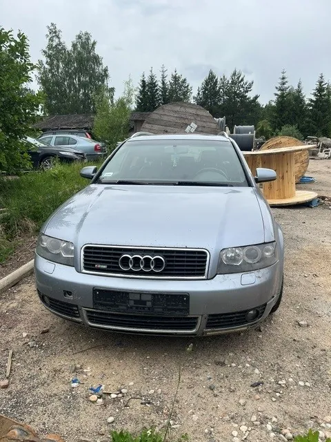 Srebrny samochód marki Audi zaparkowany na piaszczystym terenie. Widoczny jest przód pojazdu z charakterystycznym grillem i czterema pierścieniami logo Audi. W tle znajdują się zielone drzewa, krzewy oraz sterta drewnianych elementów i materiałów budowlanych.