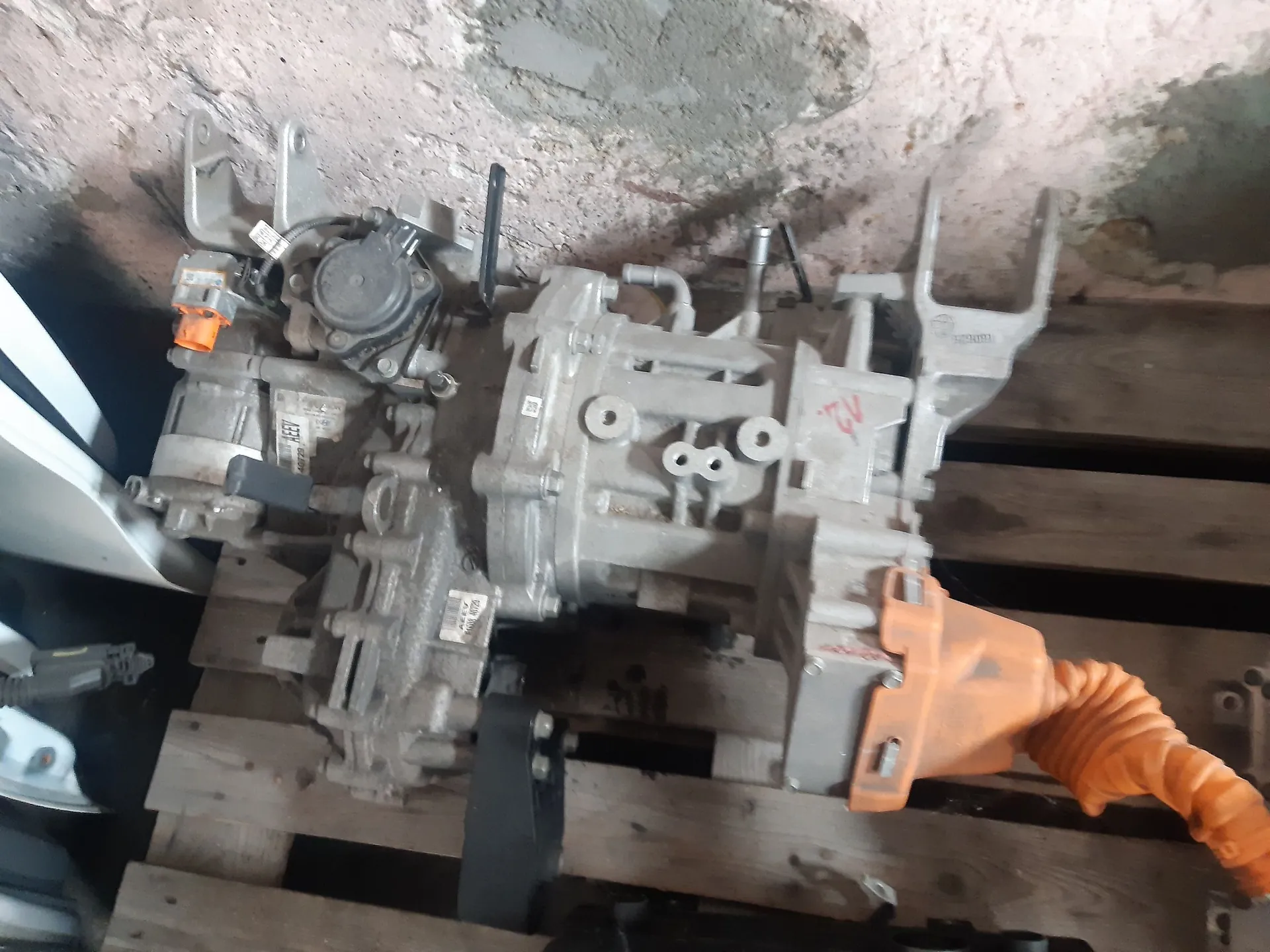 Srebrna, metalowa skrzynia biegów z samochodu, leżąca na drewnianej palecie. Element posiada widoczne złącza elektryczne, w tym jedno w kolorze pomarańczowym po prawej stronie oraz mniejsze pomarańczowe złącze po lewej. Na obudowie widoczne są odlewy metalu i śruby montażowe.