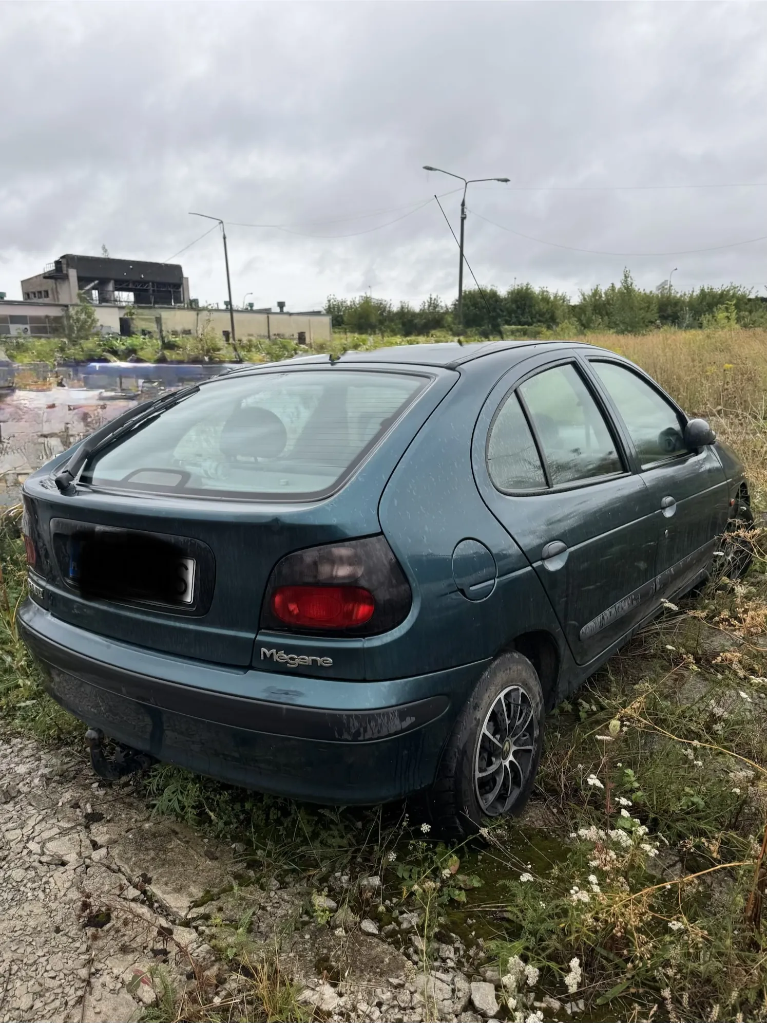 Ciemnozielony samochód marki Renault Mégane zaparkowany na nieutwardzonym terenie z trawą i kamieniami. Widoczny jest tył i prawy bok pojazdu. Na klapie bagażnika znajduje się napis „Mégane”. W tle widać szare, zachmurzone niebo, latarnie uliczne oraz fragmenty budynków przemysłowych.