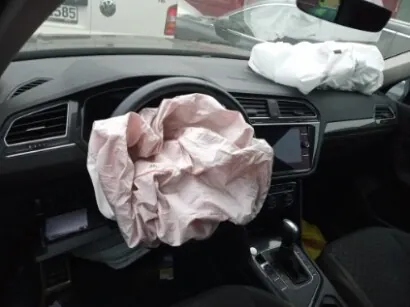 Wnętrze samochodu z rozwiniętymi poduszkami powietrznymi: różowa poduszka przed kierownicą i biała po stronie pasażera. Widoczna deska rozdzielcza z ekranem, dźwignia biegów oraz żółty przedmiot w pobliżu niej. Przez przednią szybę widoczny fragment innego pojazdu z częściowo widoczną tablicą rejestracyjną zawierającą cyfry '585'.