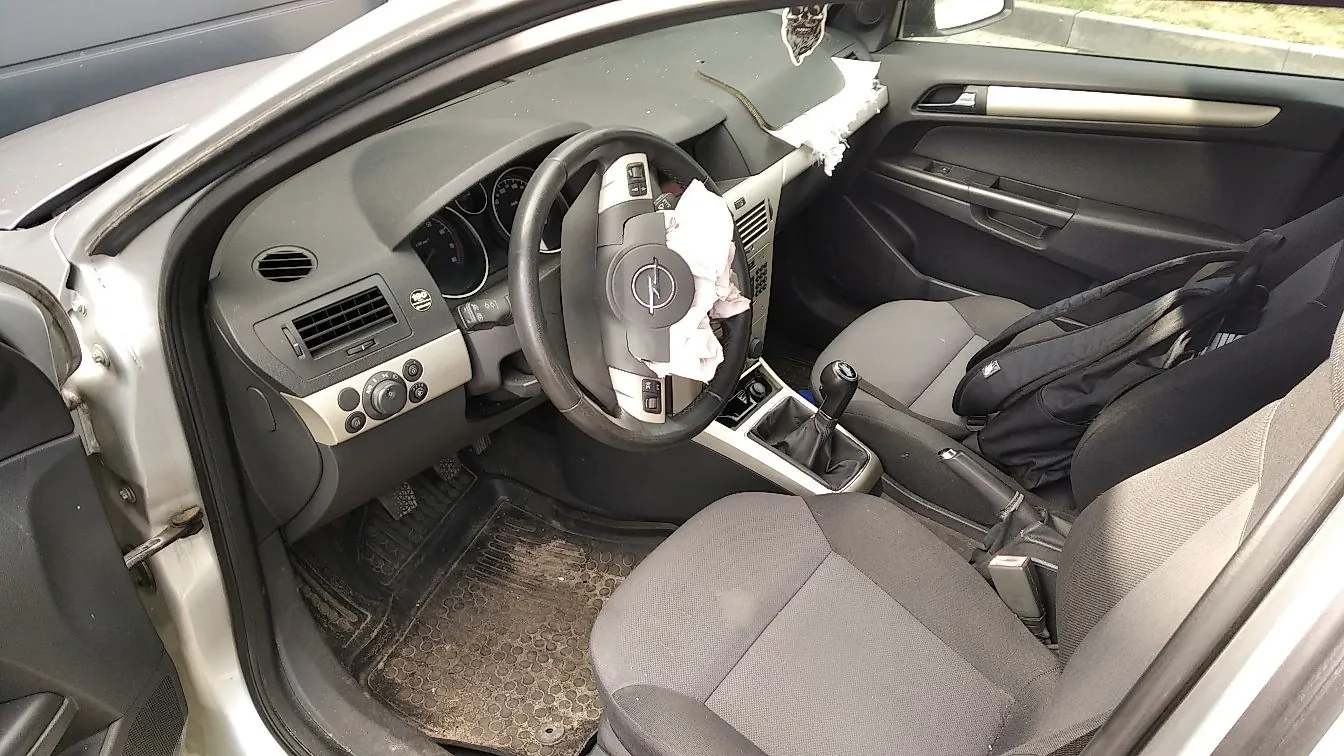 Wnętrze samochodu widziane z perspektywy otwartych drzwi kierowcy. Widoczna jest szara deska rozdzielcza, kierownica z logo marki Opel, dwa przednie szare fotele oraz lewarek zmiany biegów. Na kierownicy oraz na górnej części deski rozdzielczej znajdują się białe szmatki. Na siedzeniu pasażera leży czarna torba. Podłoga wyłożona jest gumowymi dywanikami.