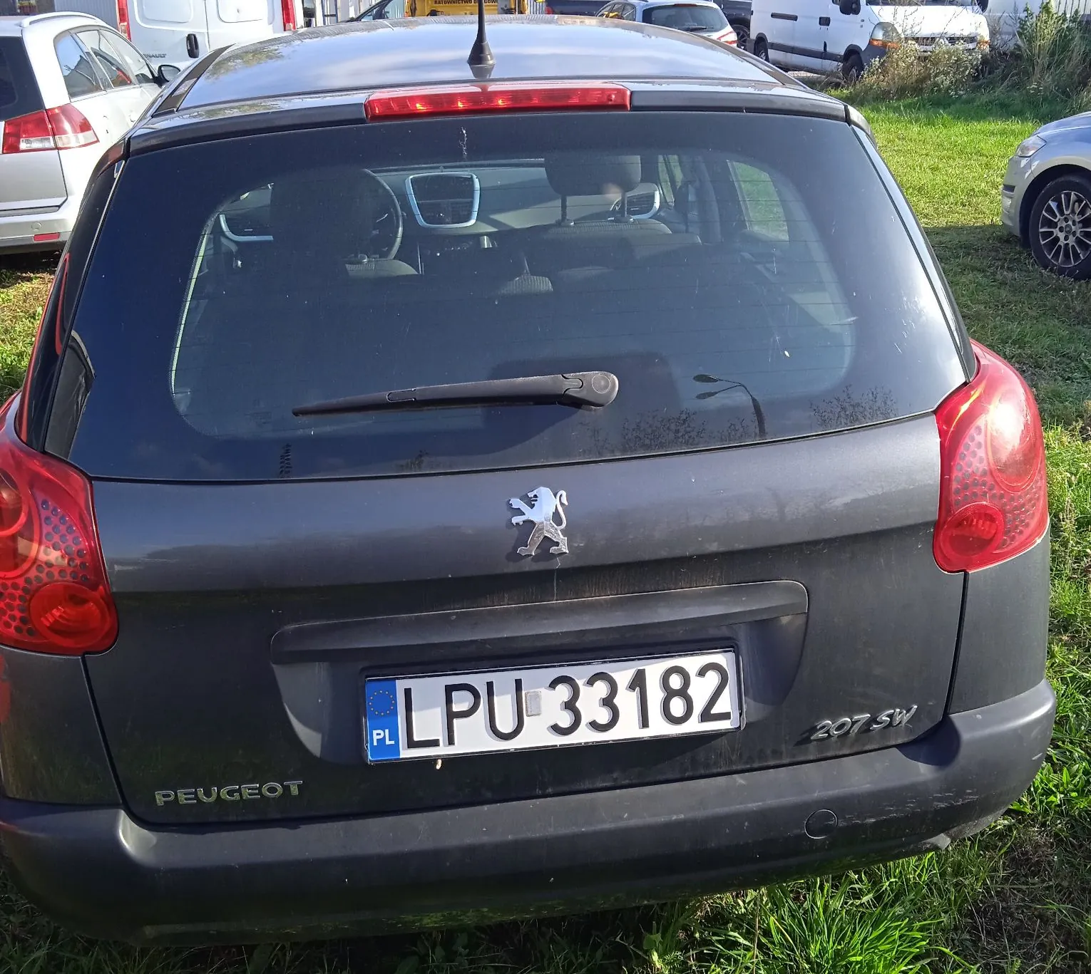 Tylny widok szarego samochodu marki Peugeot 207 SW. Na klapie bagażnika znajduje się logo lwa Peugeot oraz polska tablica rejestracyjna o numerze LPU 33182. Na zderzaku widnieje napis PEUGEOT. W tle widoczne są inne zaparkowane samochody oraz trawa.