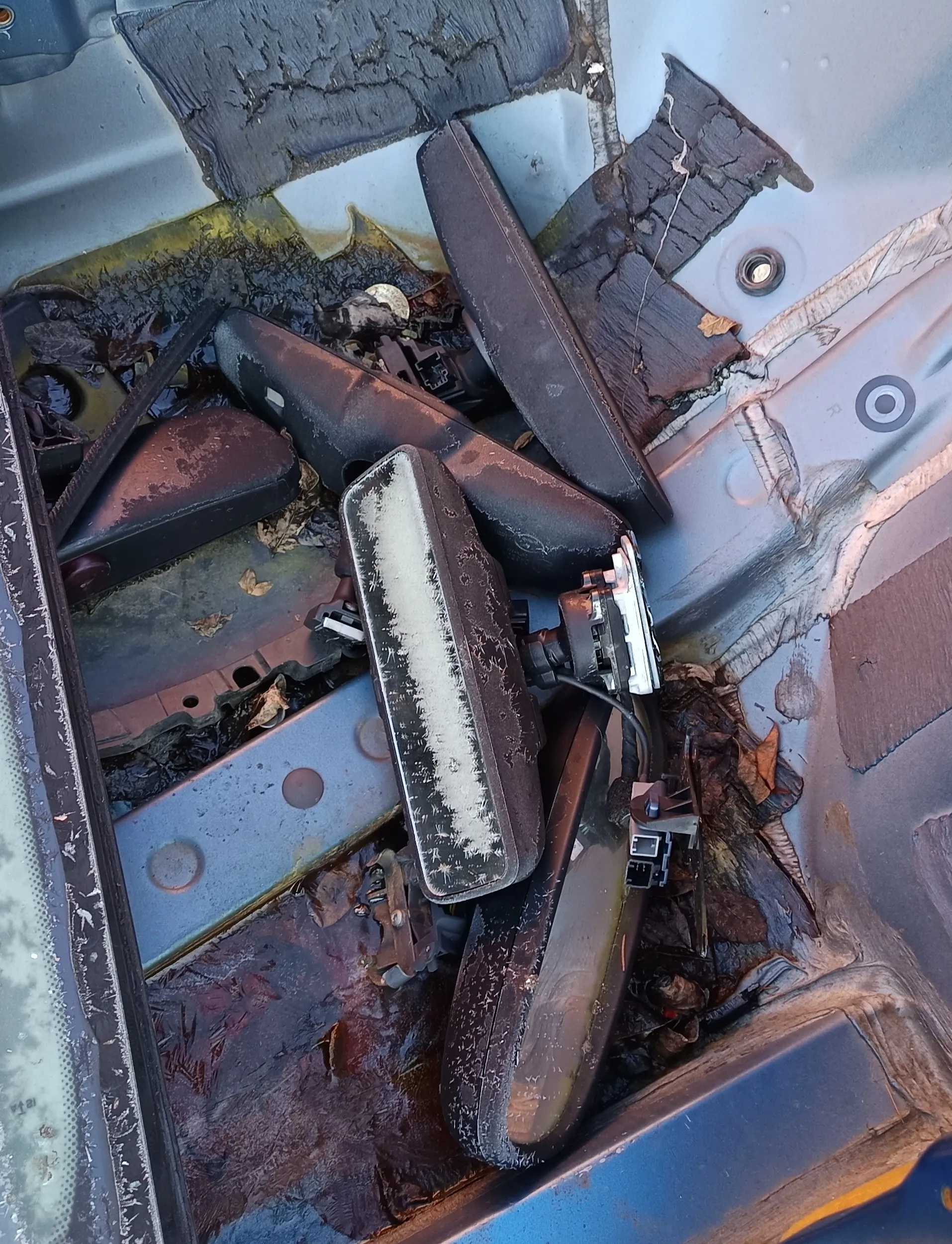 Zdjęcie przedstawia spalone i uszkodzone elementy wnętrza pojazdu. Widoczne są czarne, popękane i zdeformowane części plastikowe oraz metalowe, pokryte popiołem i rdzą. Wśród nich znajdują się elementy z białymi plamami, przewody elektryczne oraz suchymi liśćmi. Tło stanowi metalowa powierzchnia z pęknięciami, plamami i śladami korozji. Całość pokryta jest warstwą brudu i spalenizny.