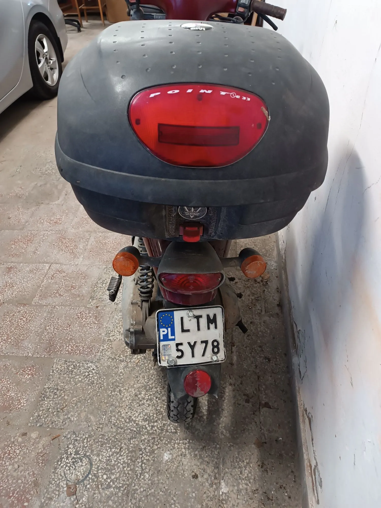 Widok z tyłu na skuter z zamontowanym dużym, czarnym kufrem bagażowym. Na kufrze znajduje się czerwone światło stopu oraz napis 'POINT'. Poniżej widoczna jest tablica rejestracyjna o numerze LTM 5Y78 z oznaczeniem PL. Skuter stoi na jasnej, kamiennej posadzce obok białej ściany.