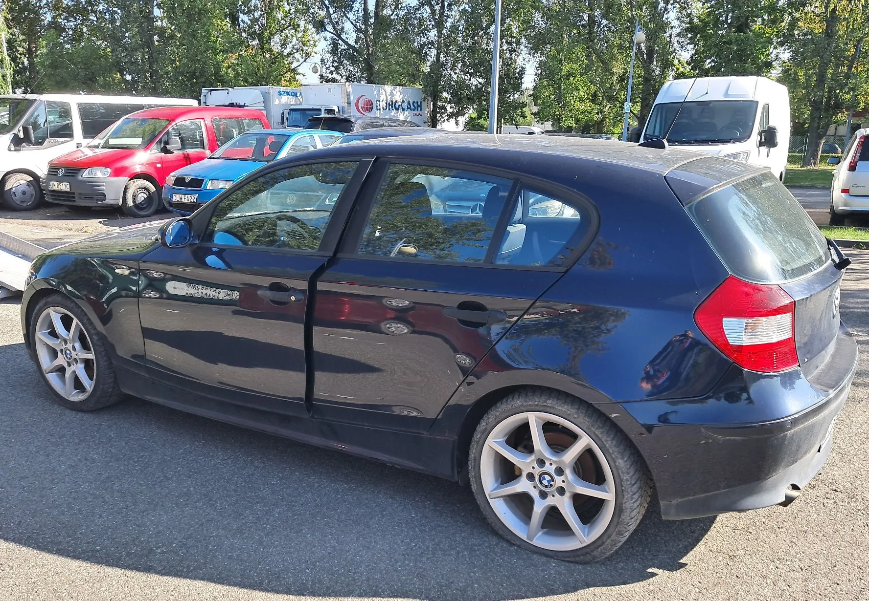 Czarny samochód osobowy marki BMW w nadwoziu hatchback zaparkowany na asfaltowym placu. W tle widoczne są inne pojazdy: czerwony samochód dostawczy, niebieski samochód oraz białe vany. W oddali znajdują się zielone drzewa i biała przyczepa z napisem 'EUROCASH'.